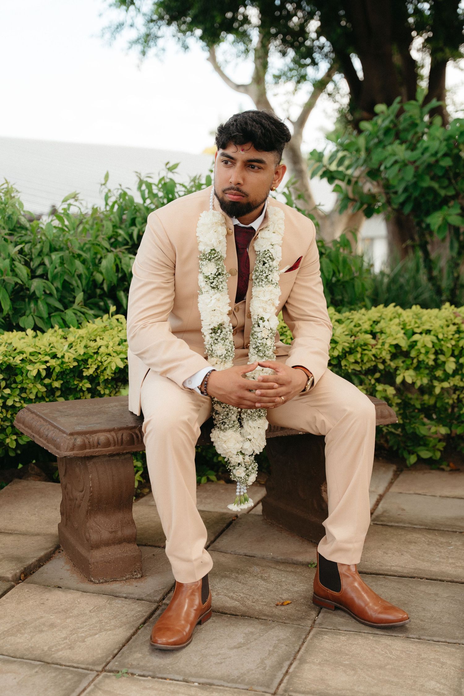 A person in a beige suit and floral garland sits on a stone bench in a garden setting.