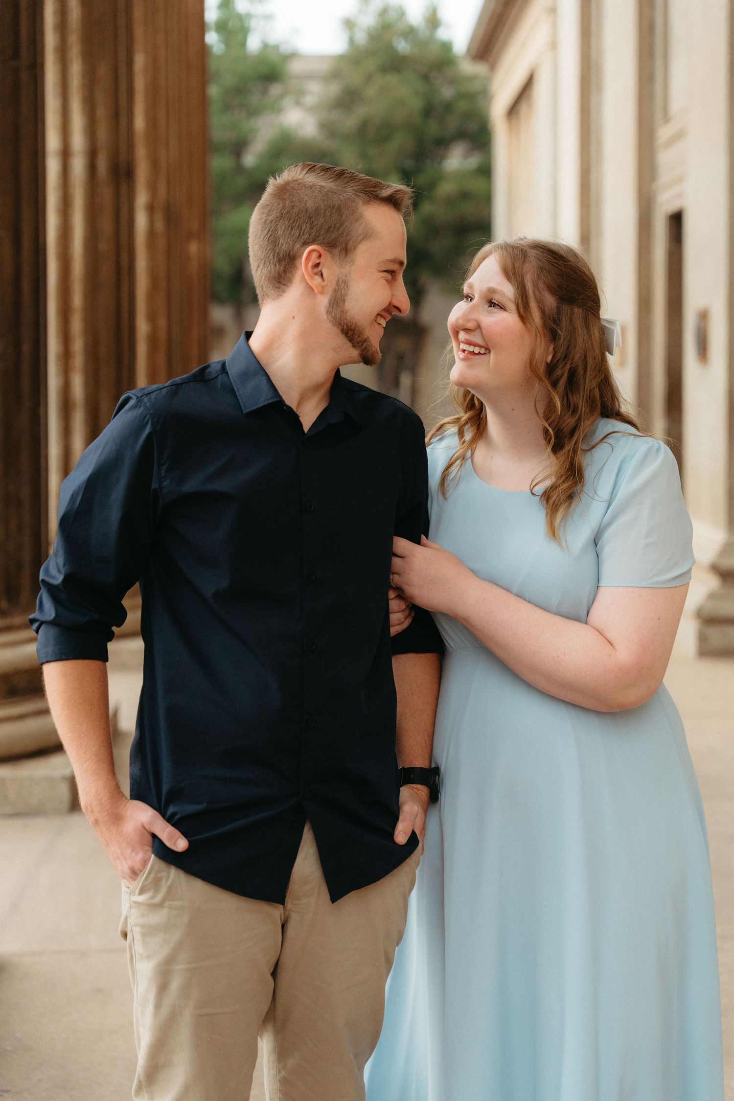 A couple in blue dress and navy shirt share loving glances while posing in an outdoor colonnade setting.