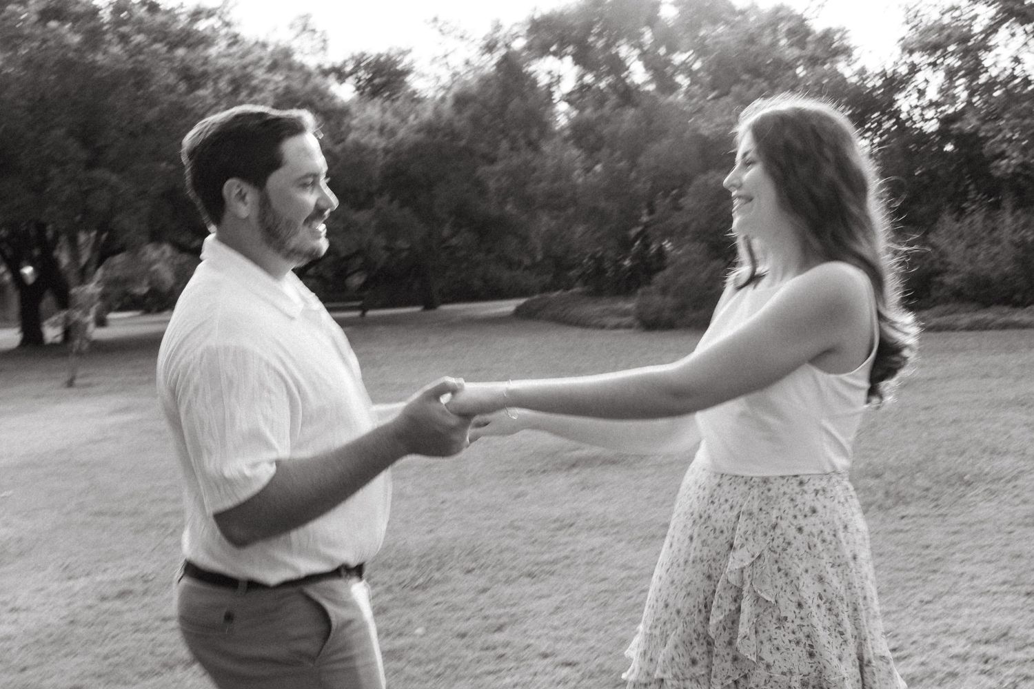 Black and white photo of a smiling couple holding hands and dancing outdoors in a park setting.