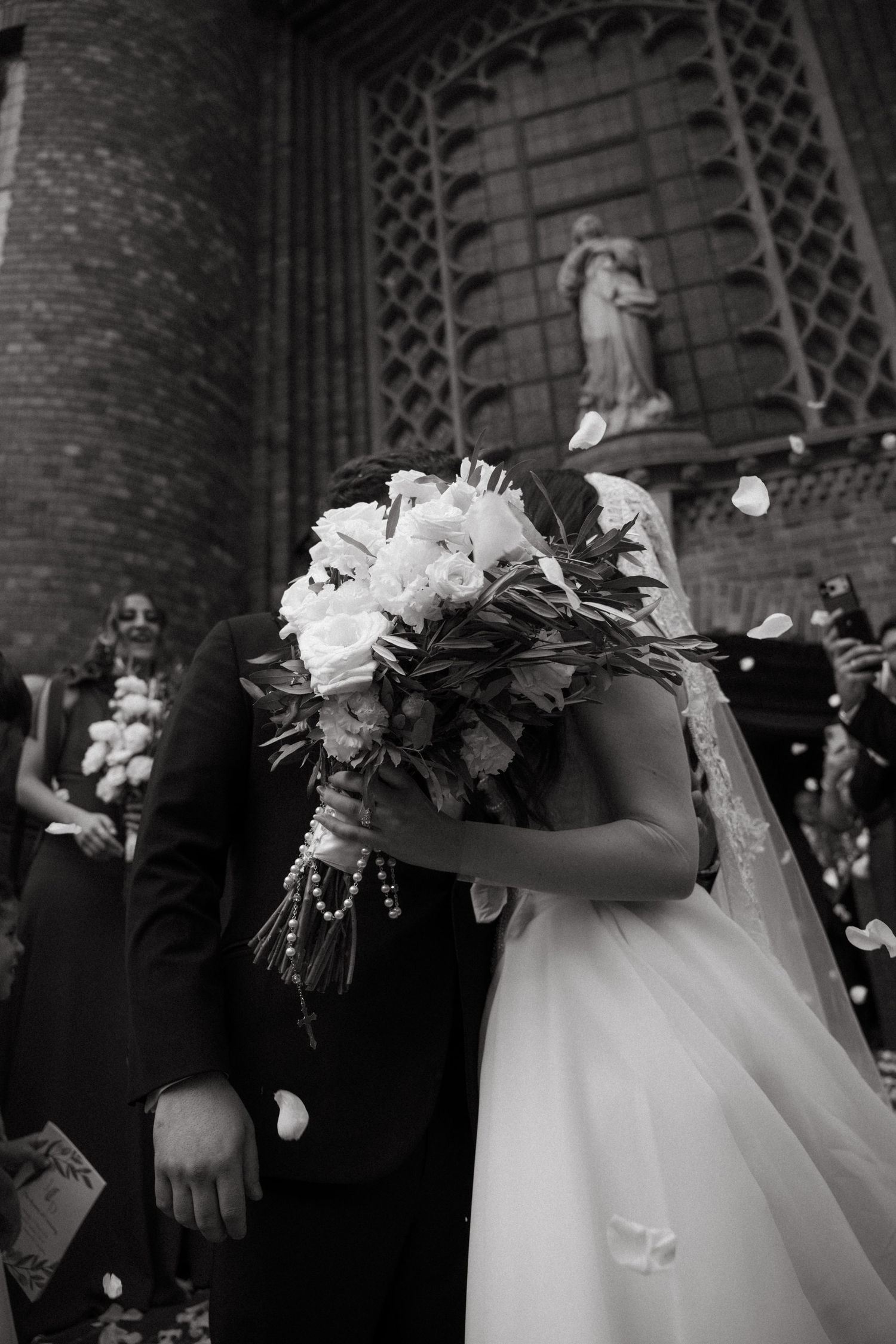 Black and white photo of bride and groom kissing behind white rose bouquet outside Rosebank Catholic Church