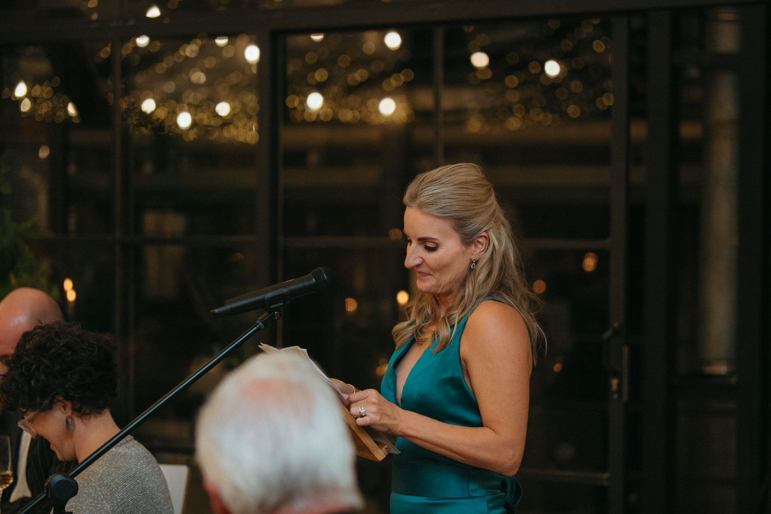 Woman in teal dress speaking at podium with string lights backdrop at elegant indoor event.
