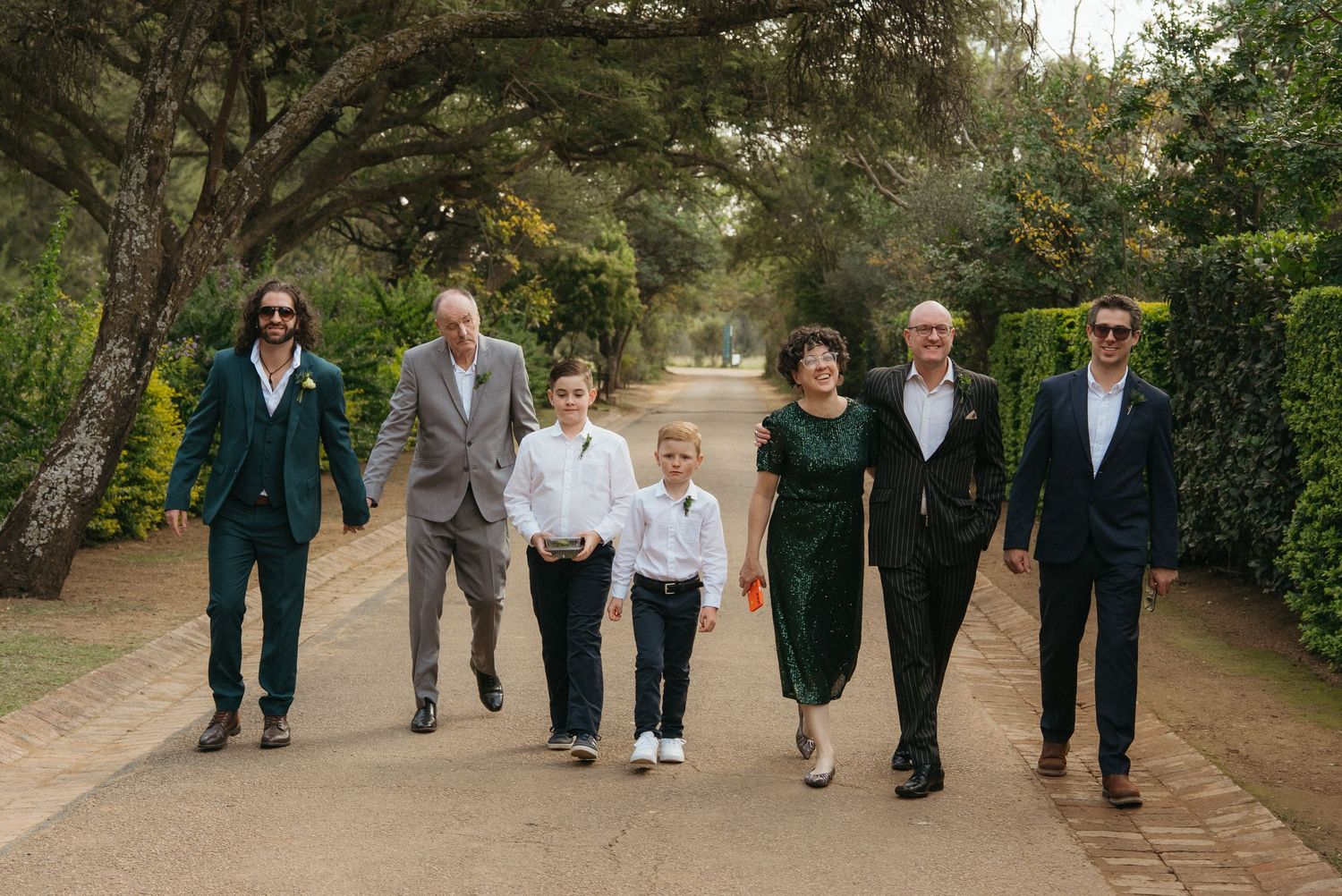 Group of seven people walking together on a tree-lined path, dressed in formal attire for a wedding.