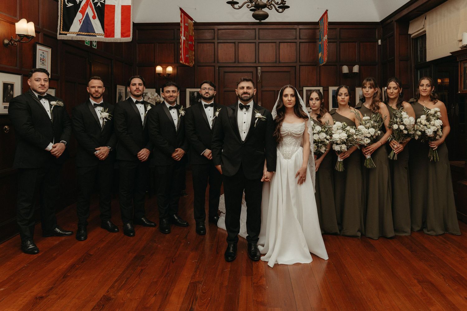 Wedding party poses in wood-paneled hall Glenshiel, bride in white gown with groomsmen in black tuxedos and bridesmaids in olive dresses.