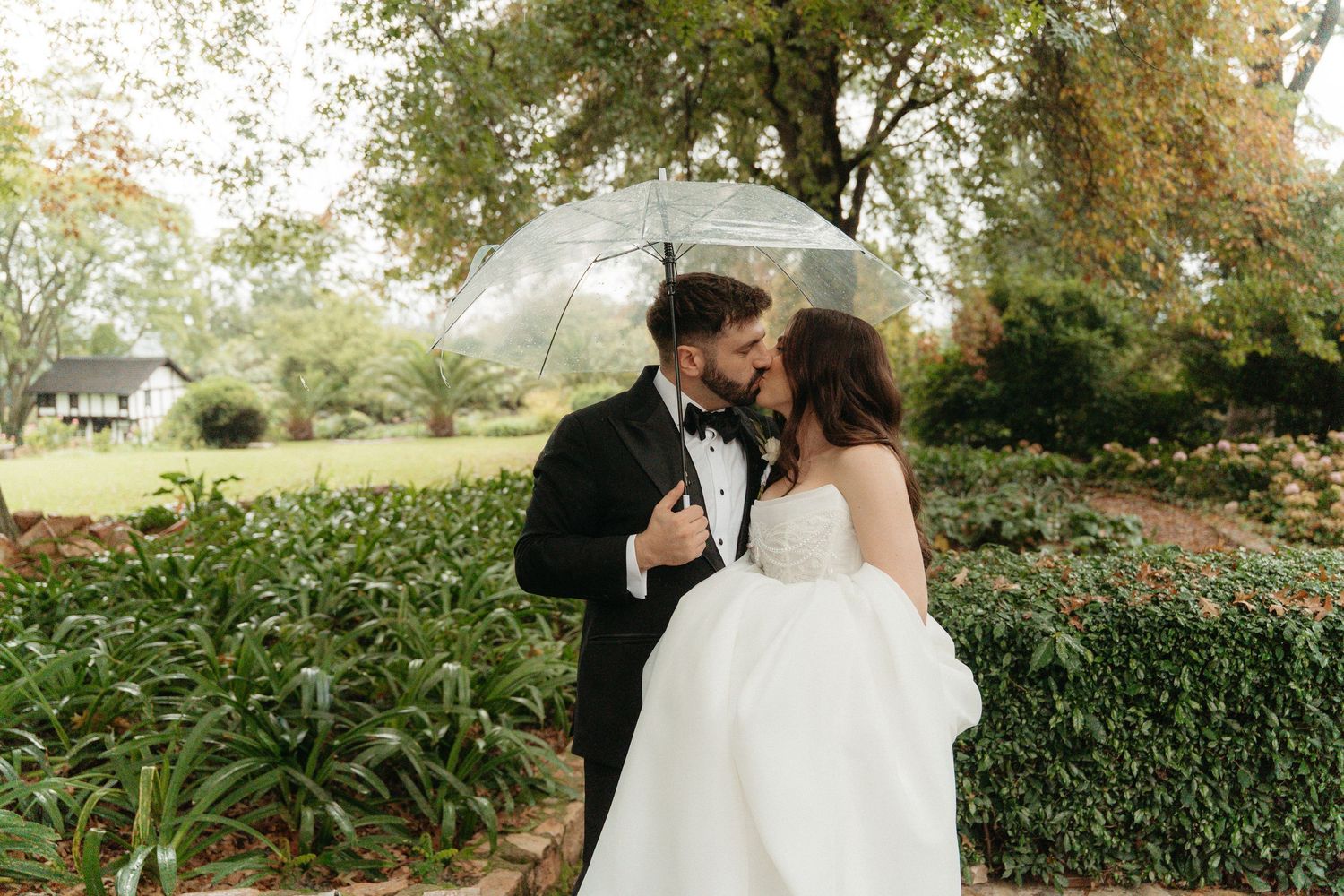 Bride and groom share a romantic kiss under a clear umbrella in a lush garden on their wedding day.