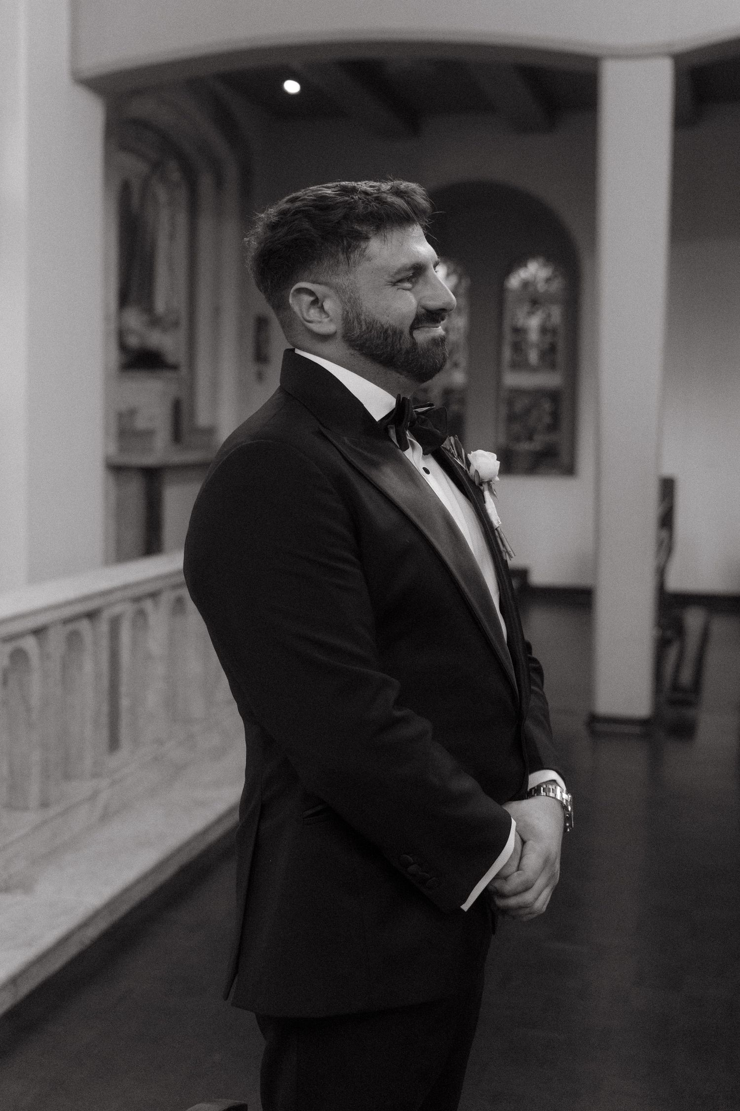 Black and white photo of a smiling groom in a tuxedo with bow tie and boutonniere standing in a church.