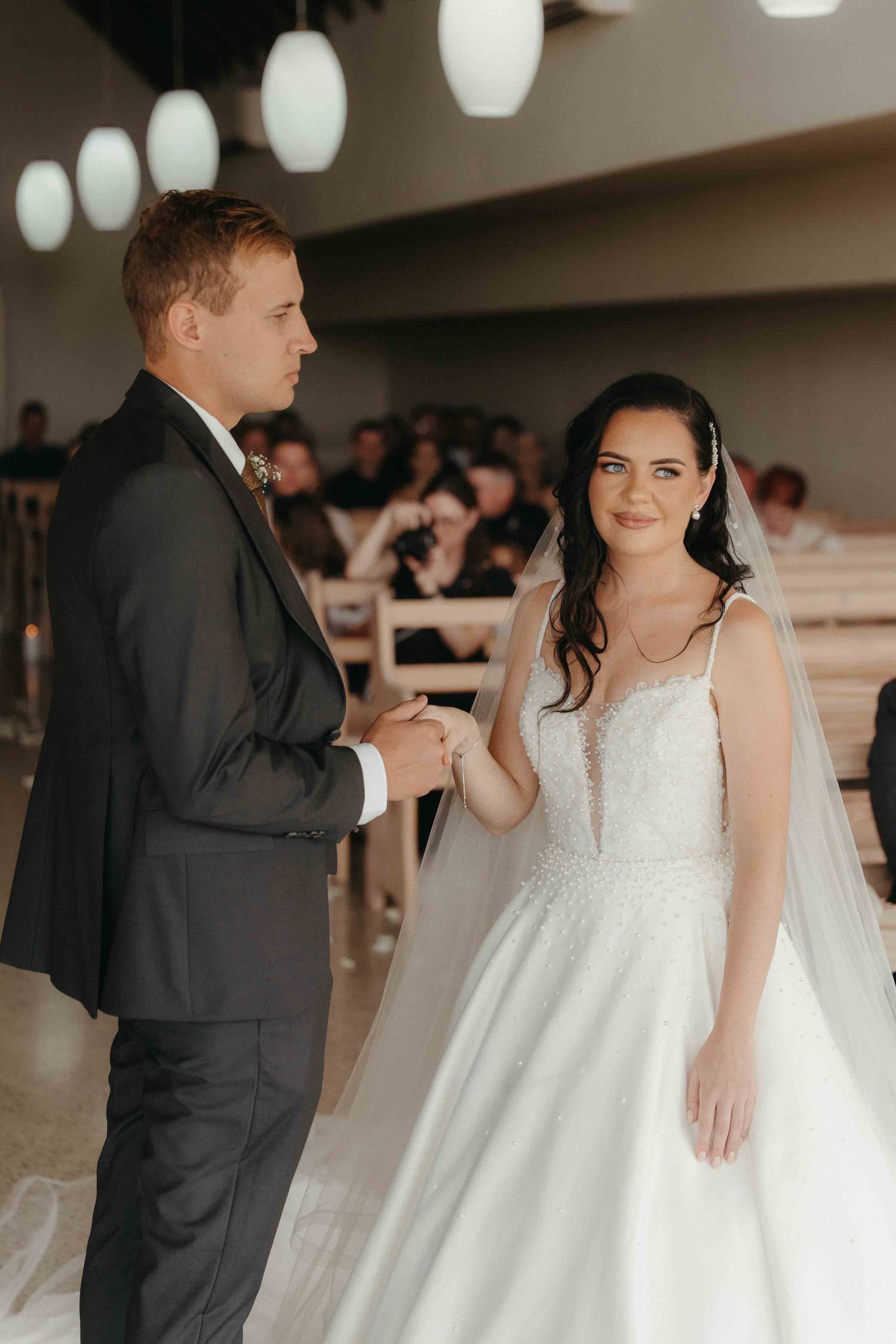Bride in white gown and groom in dark suit exchange vows at church altar, holding hands during wedding ceremony.