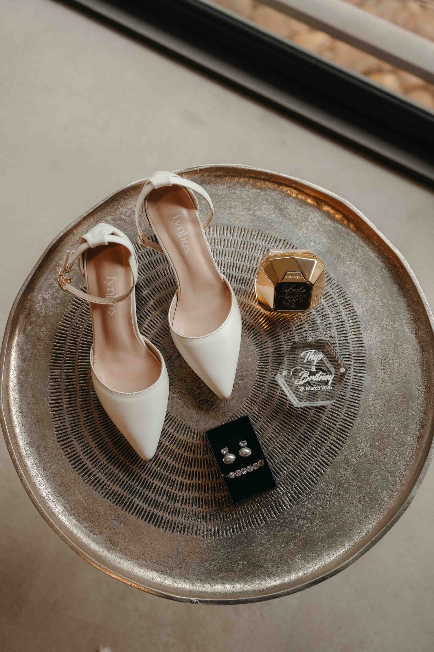 White bridal heels with ankle strap displayed on silver tray with pearl earrings and gold hexagon perfume bottle.