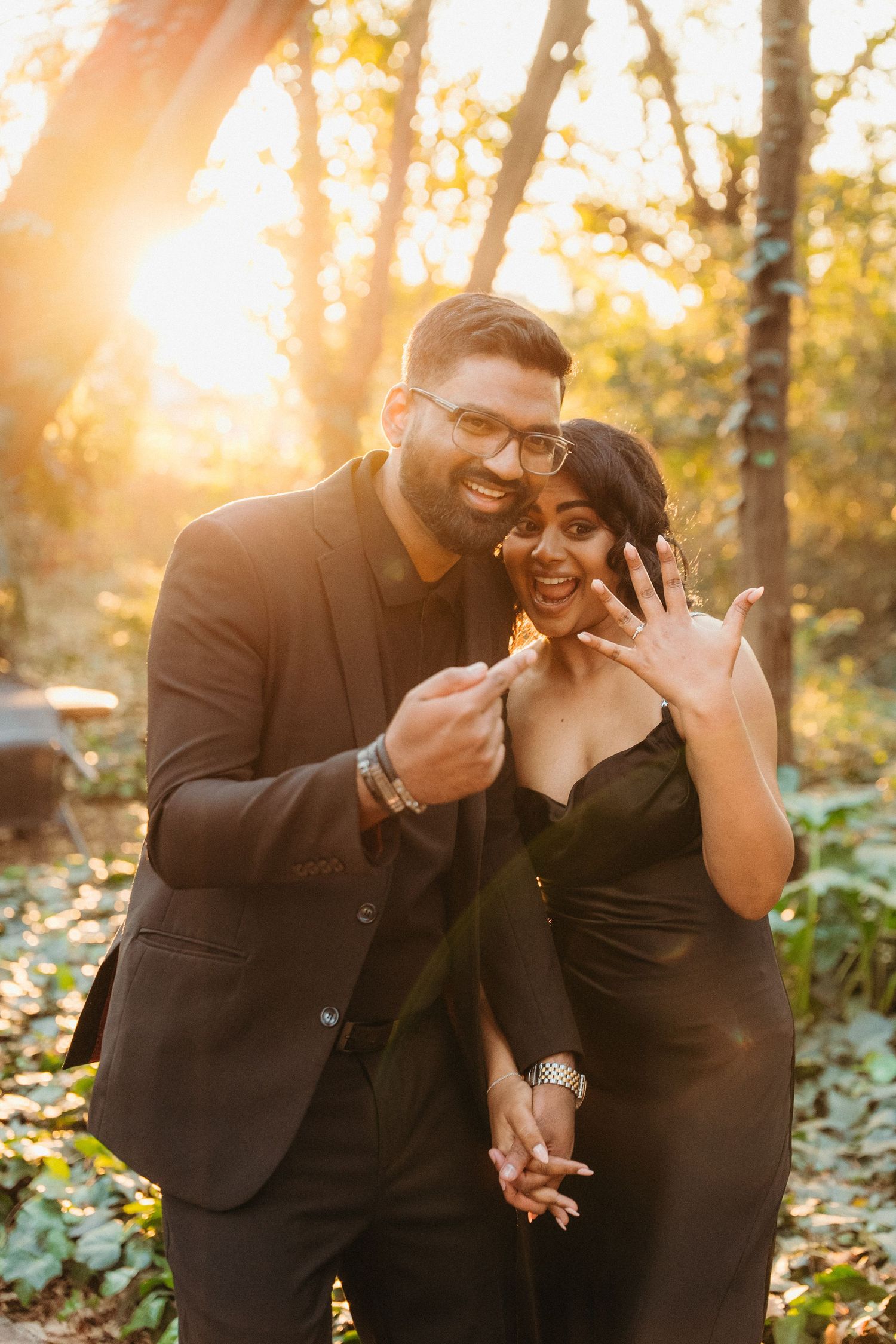 Couple in black outfits showing engagement ring outdoors in golden sunset light among trees.