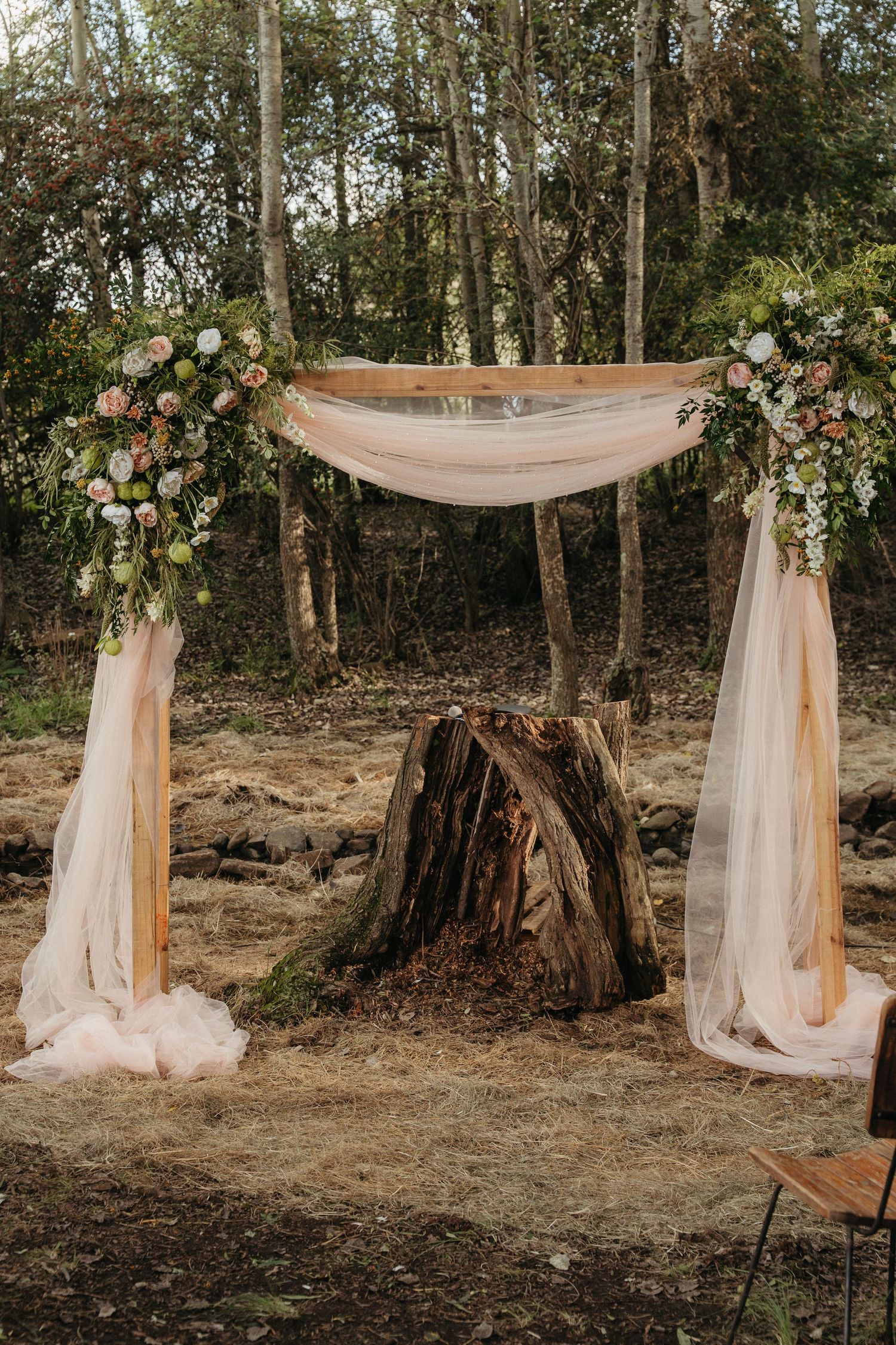 Rustic outdoor wedding arch with blush draping, floral arrangements, and a tree stump centerpiece in a forest setting.