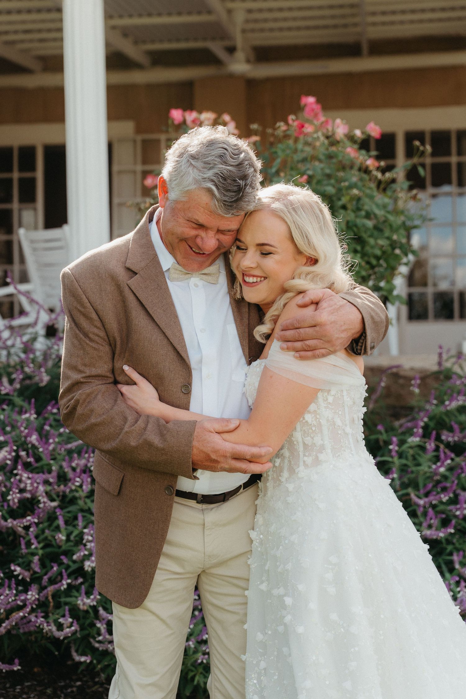 A bride in a white floral gown embraces an older man in a brown blazer outdoors near blooming flowers.