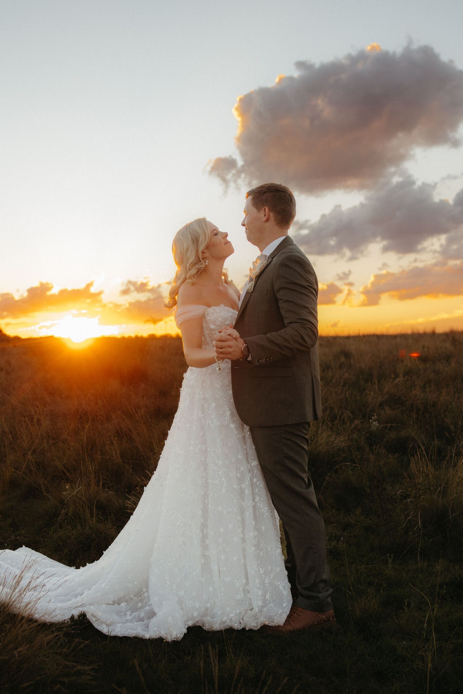 A bride and groom share a romantic moment at sunset in a field, with golden light and dramatic clouds behind them.