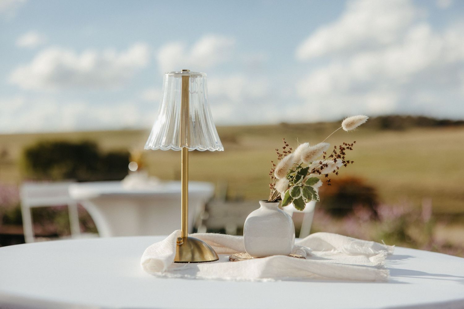 Elegant outdoor table setting with a gold lamp and white ceramic vases on a white table with a scenic field backdrop.