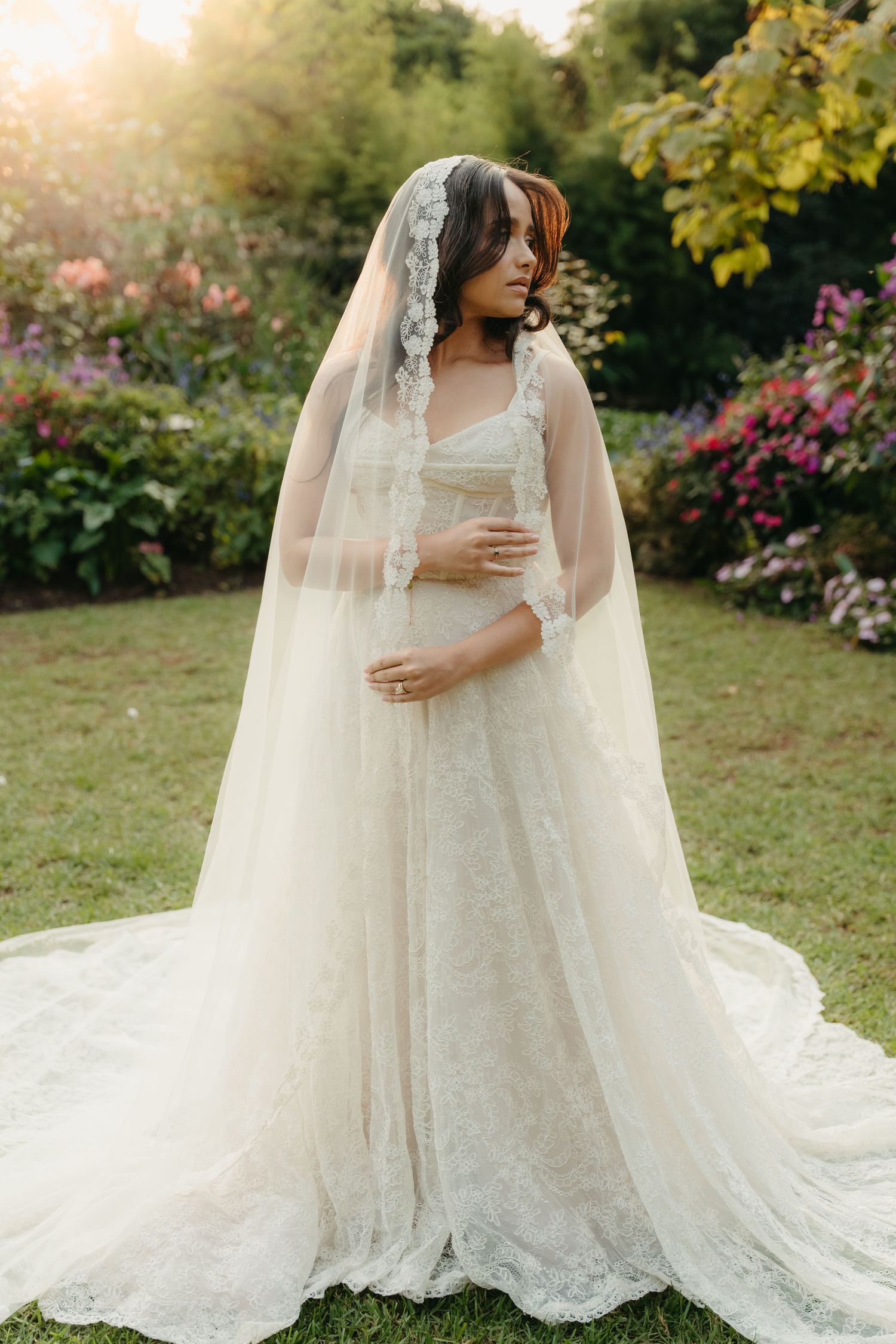 Bride in lace wedding gown with cathedral veil stands in sunlit Beechwood garden with colorful flowers.