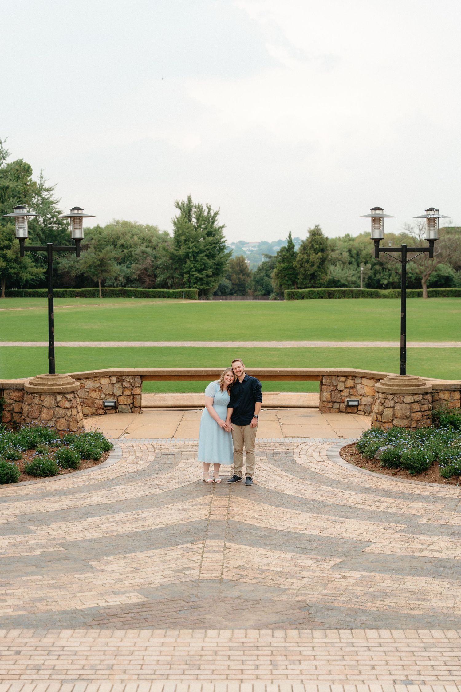 A couple stands on a paved stone terrace overlooking a green lawn with lampposts and trees in the background.