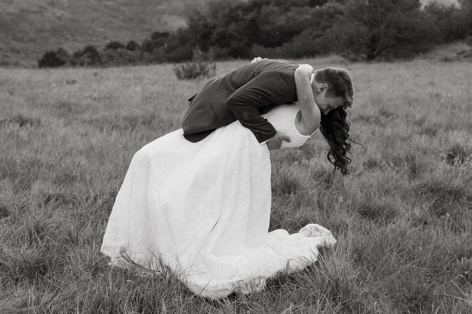 A couple in wedding attire sits together on a grassy hillside in a romantic black and white photograph.