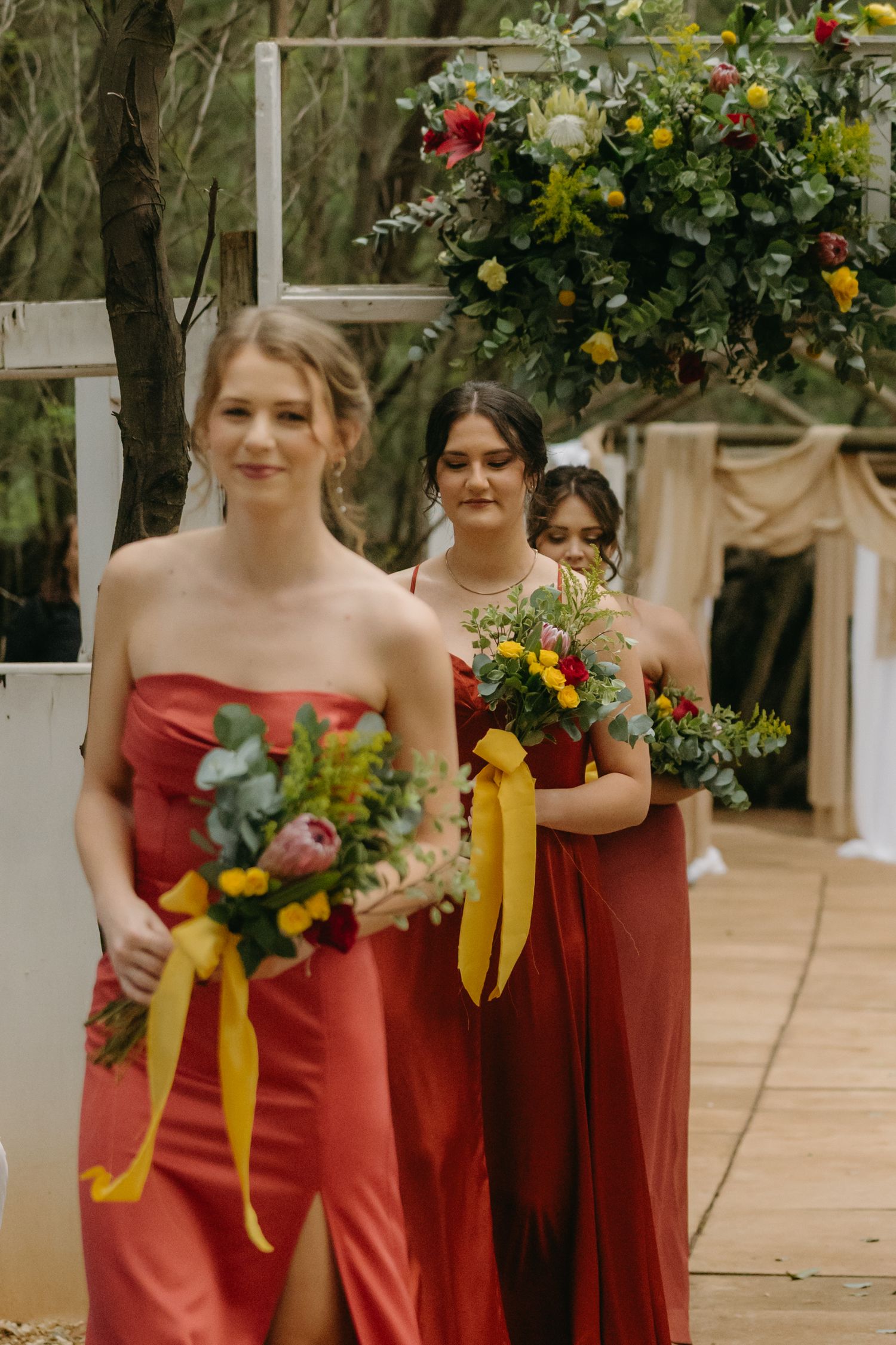 Three bridesmaids in coral-colored dresses hold bright bouquets with yellow ribbons during an outdoor ceremony.