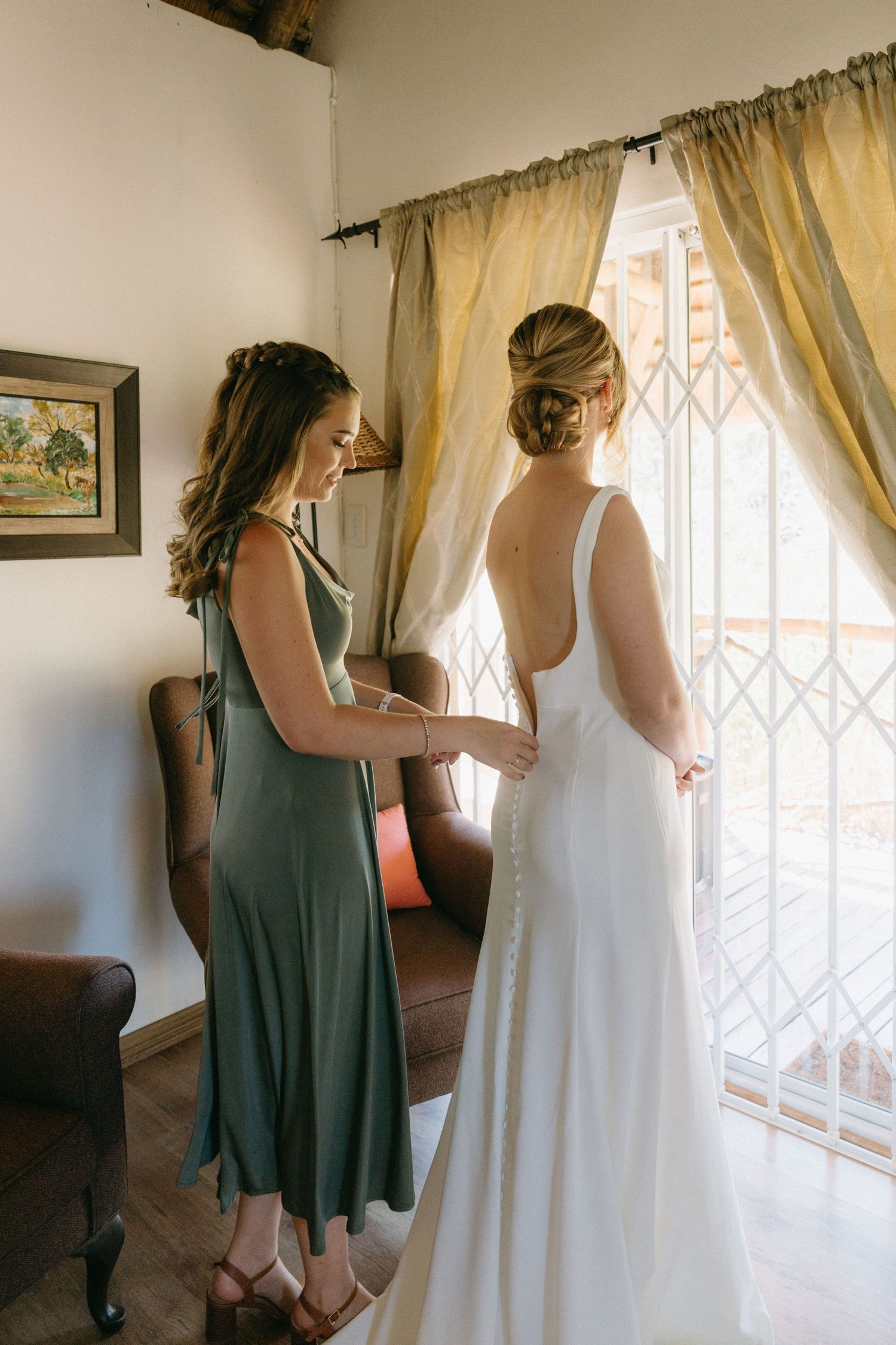 Two women in a sunlit room as one in a sage green dress helps adjust a white wedding gown.