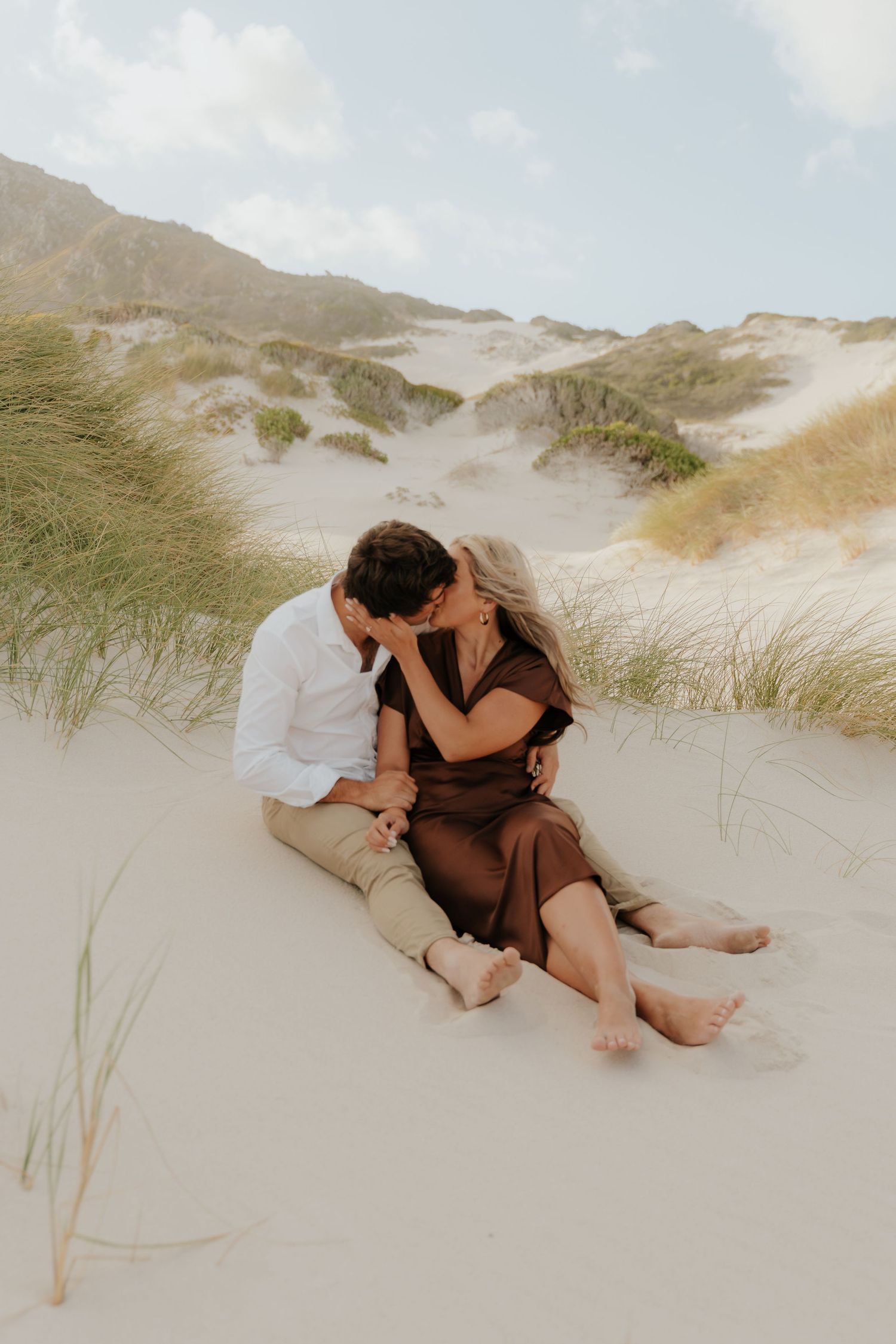 Romantic couple kissing on white sand dunes surrounded by beach grass on a sunny day.