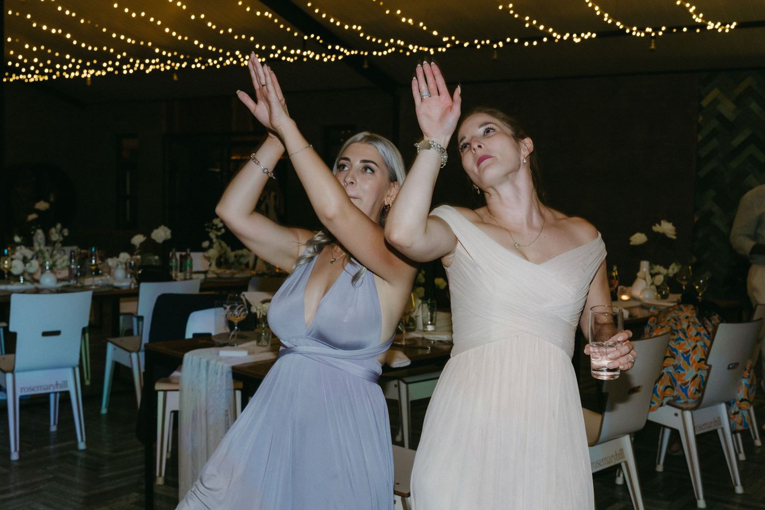 Two women in formal dresses dance together at a reception under twinkling string lights against a dark backdrop.