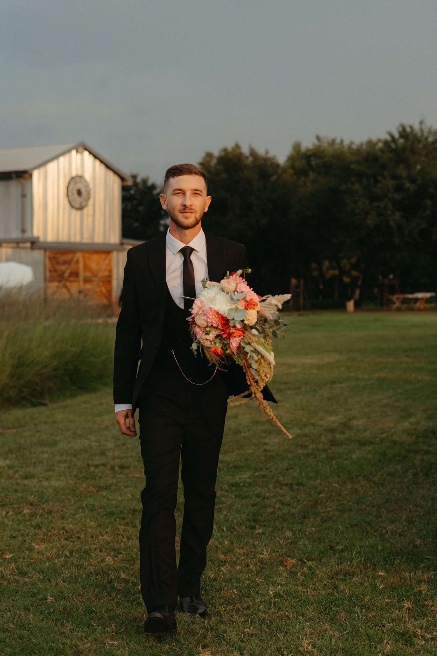 Person in dark suit holds colorful wedding bouquet while standing on grass with barn in background.