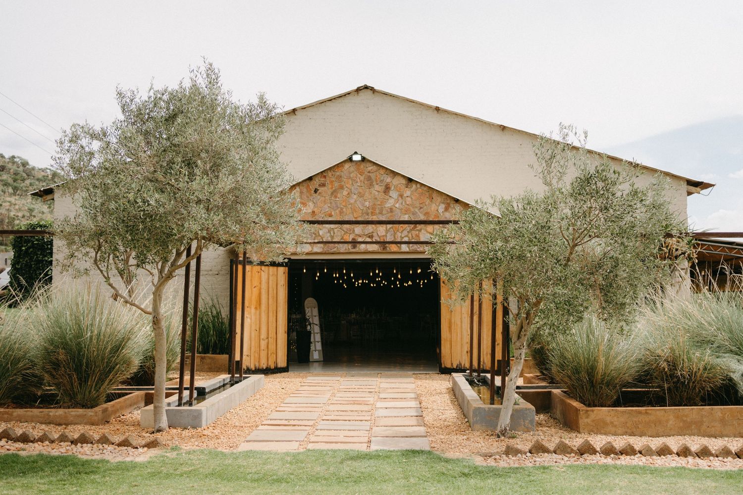Modern barn-style venue entrance with wooden doors flanked by olive trees and desert plants along a stone walkway.