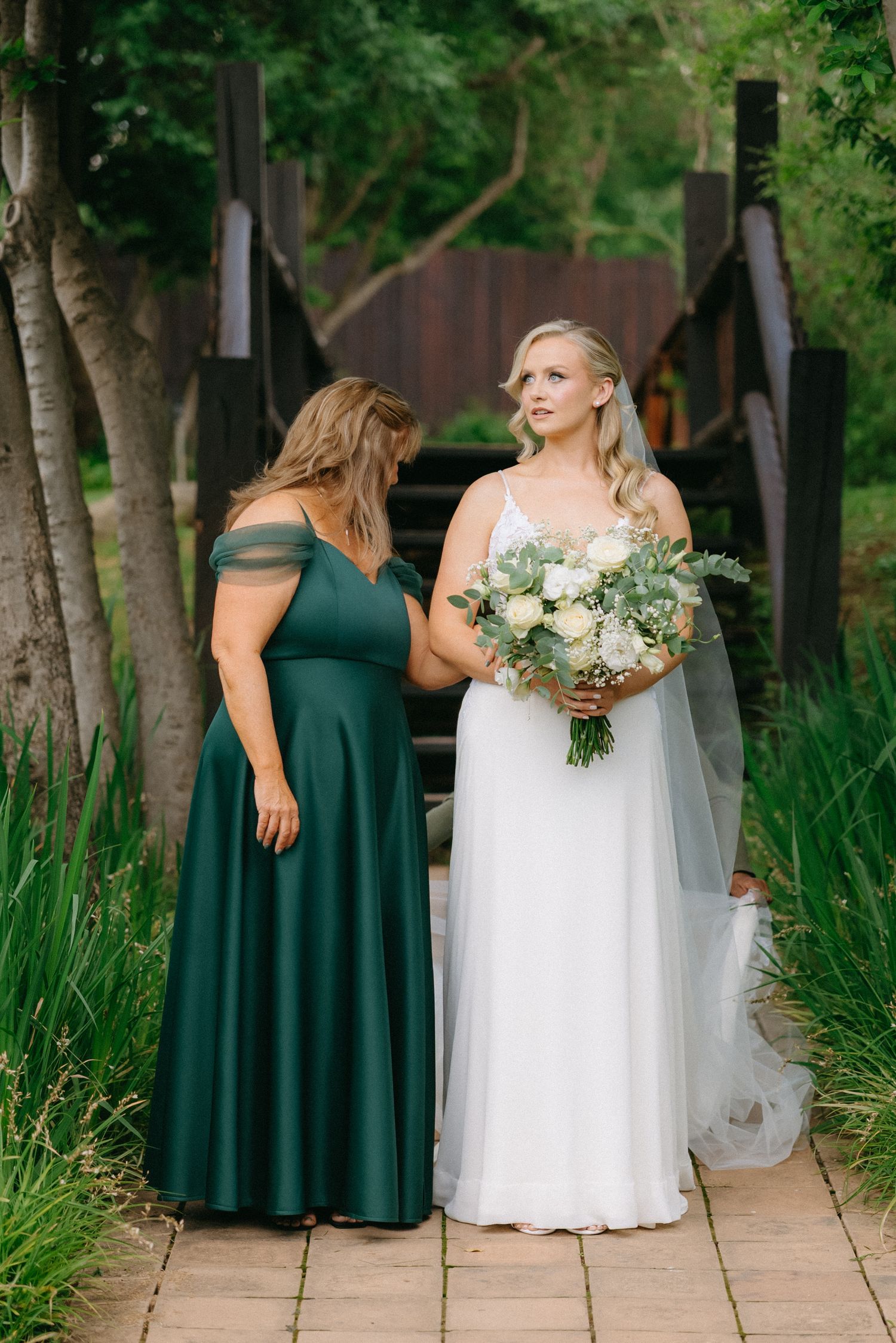 Wedding party members in emerald and white dresses stand on a garden path surrounded by greenery.