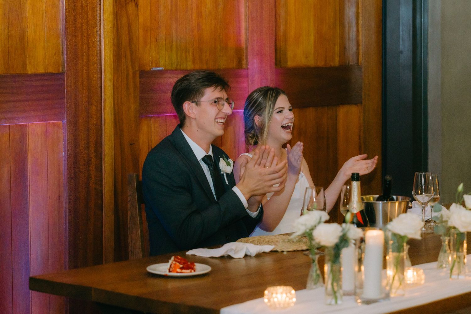 Two people laugh and react emotionally while seated at an elegant candlelit dinner table with wine glasses.
