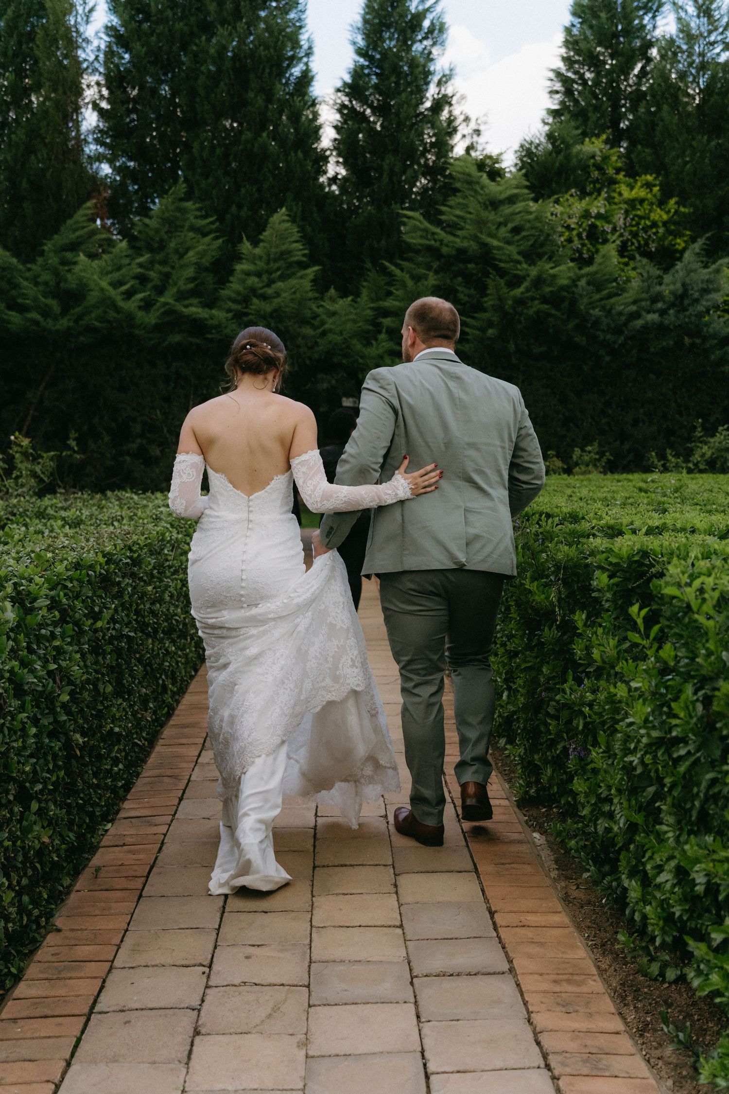 A couple walks down a brick path between manicured hedges, the bride in white and groom in grey.