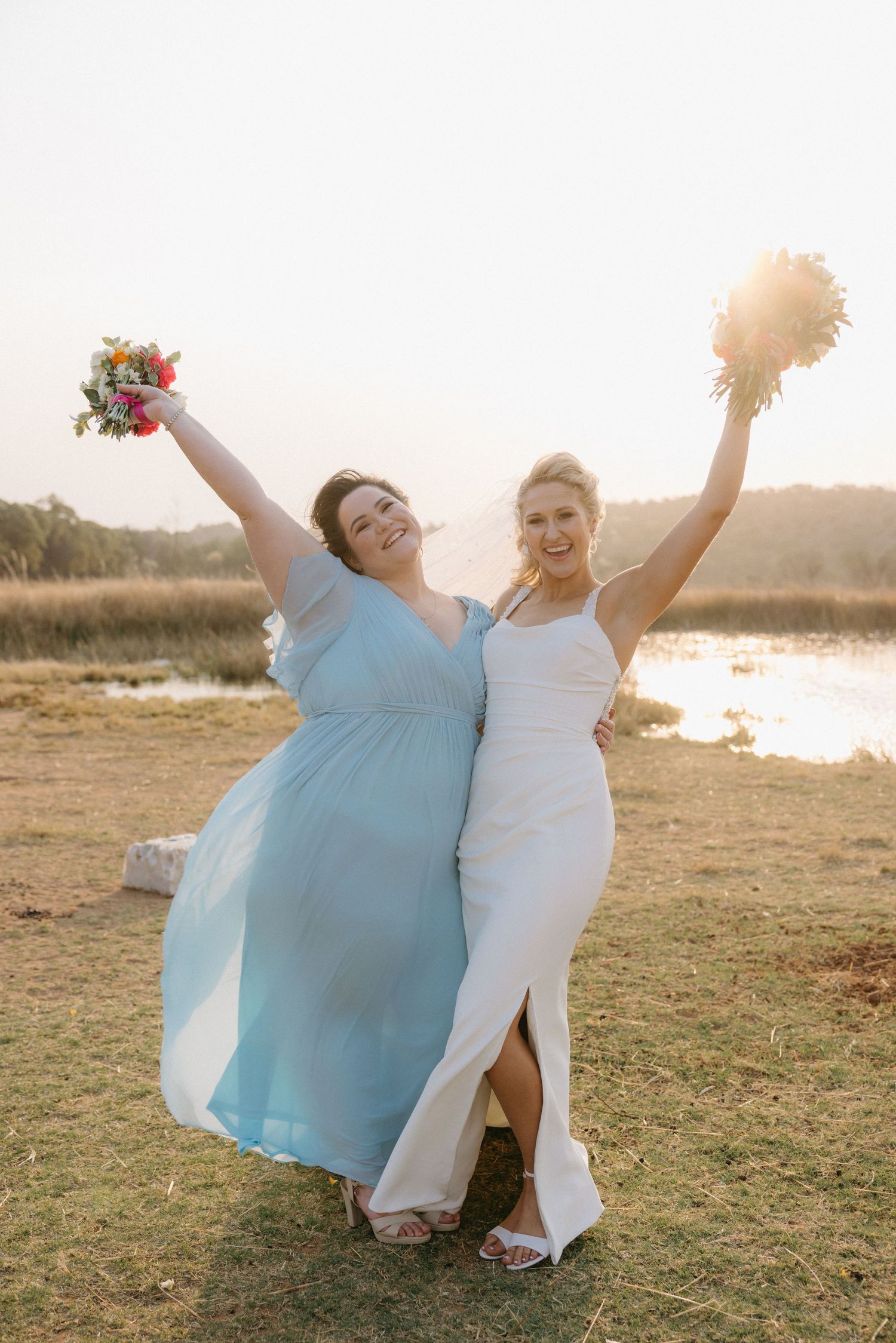 Wedding party members in light blue dresses dance joyfully at sunset near a lake.