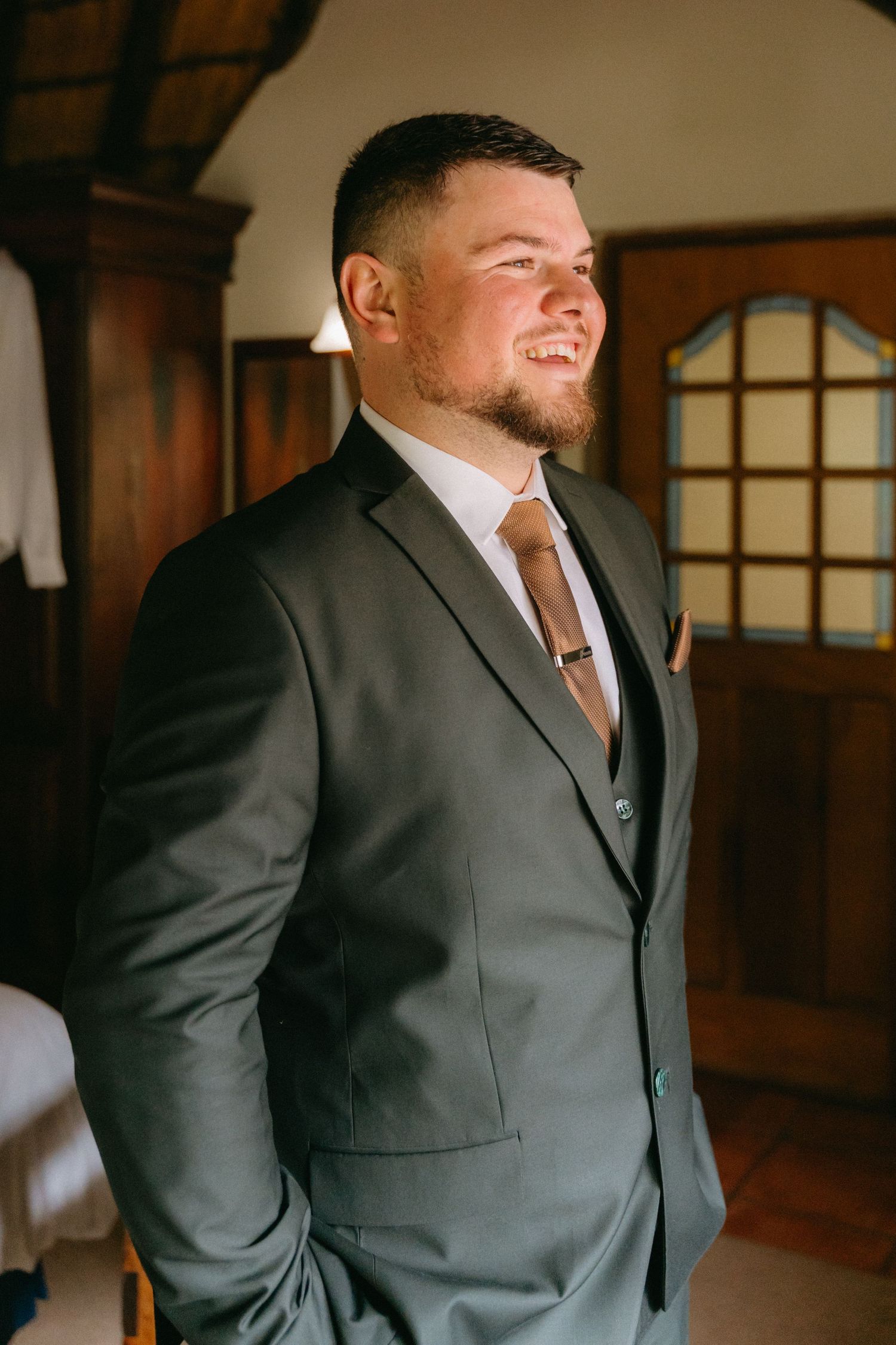 Groom in dark suit smiles while getting ready in wood-paneled room.