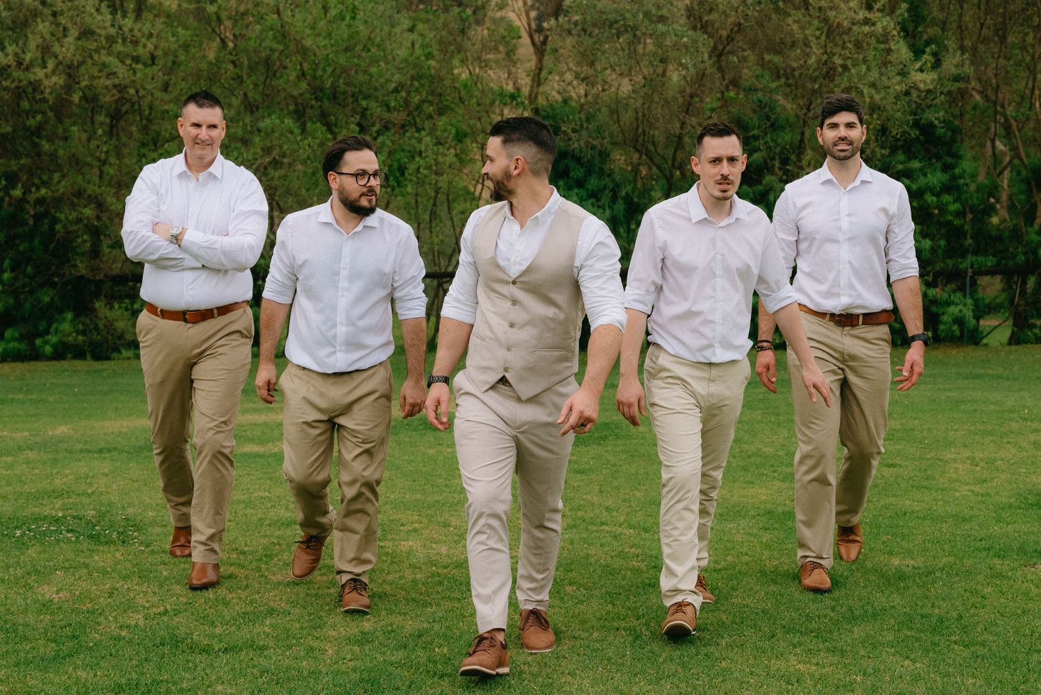 Groomsmen walking and laughing together outdoors wearing khaki pants and white shirts in a casual wedding setting.