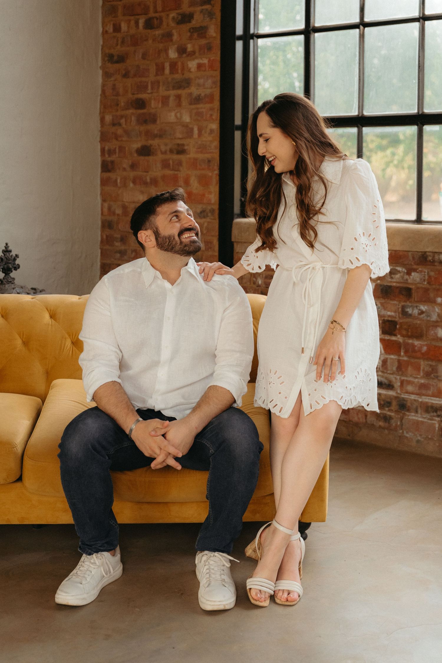 Couple in white outfits sharing intimate moments on a yellow couch in an industrial brick-walled space.