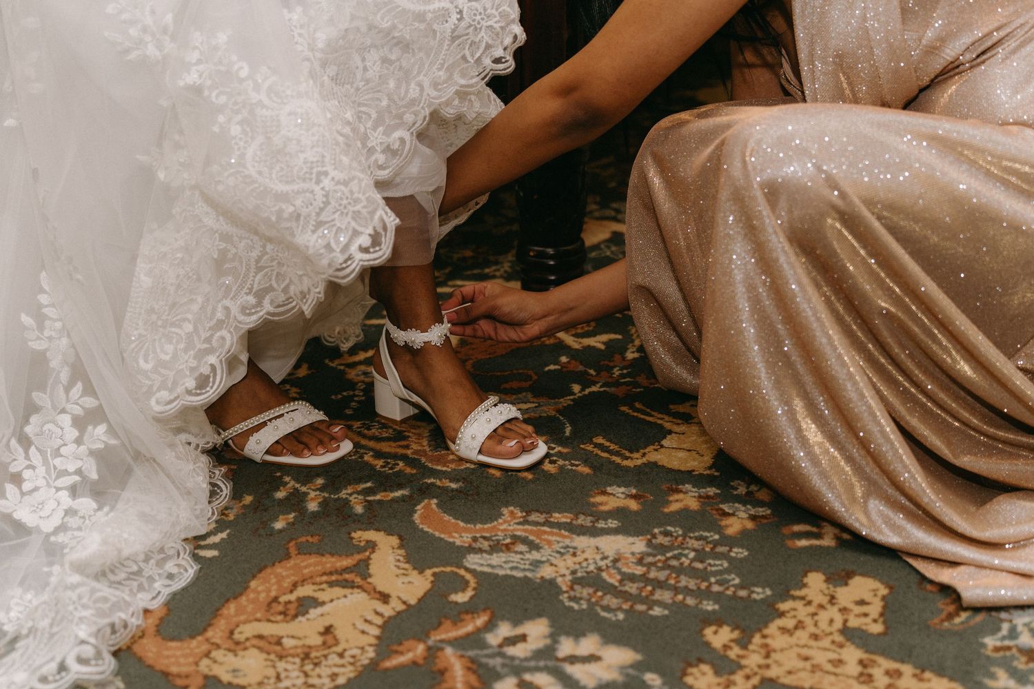 Someone helping with bridal shoes on ornate carpet during wedding preparations.