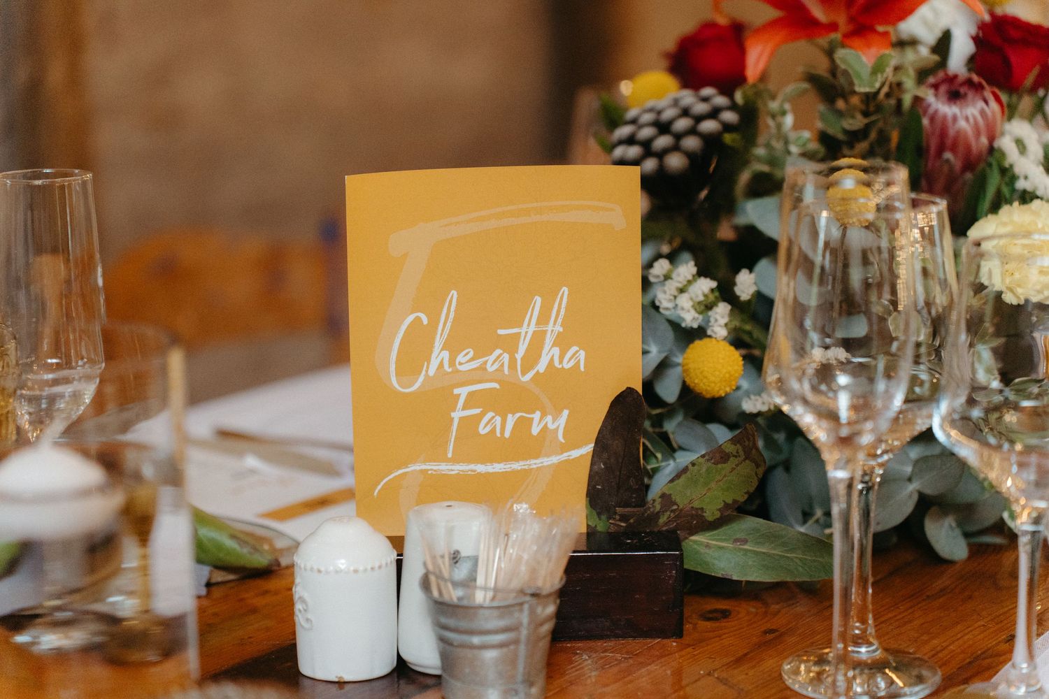 Table setting with a yellow Cheatin Farm sign, wine glasses, and floral centerpiece at a rustic event.