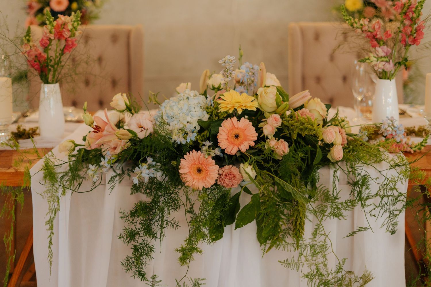 Elegant floral arrangement with peach gerbera daisies and greenery decorates a white-draped wedding head table @Boshoek Weddings, Mpumalanga