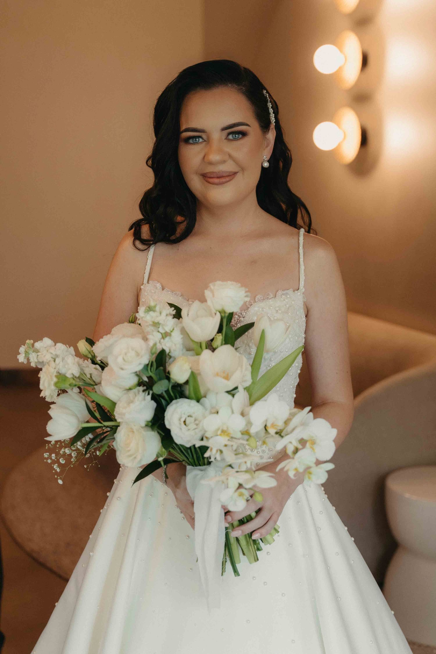 Smiling bride in white gown holding elegant white floral bouquet with roses, posing in a warmly lit room.