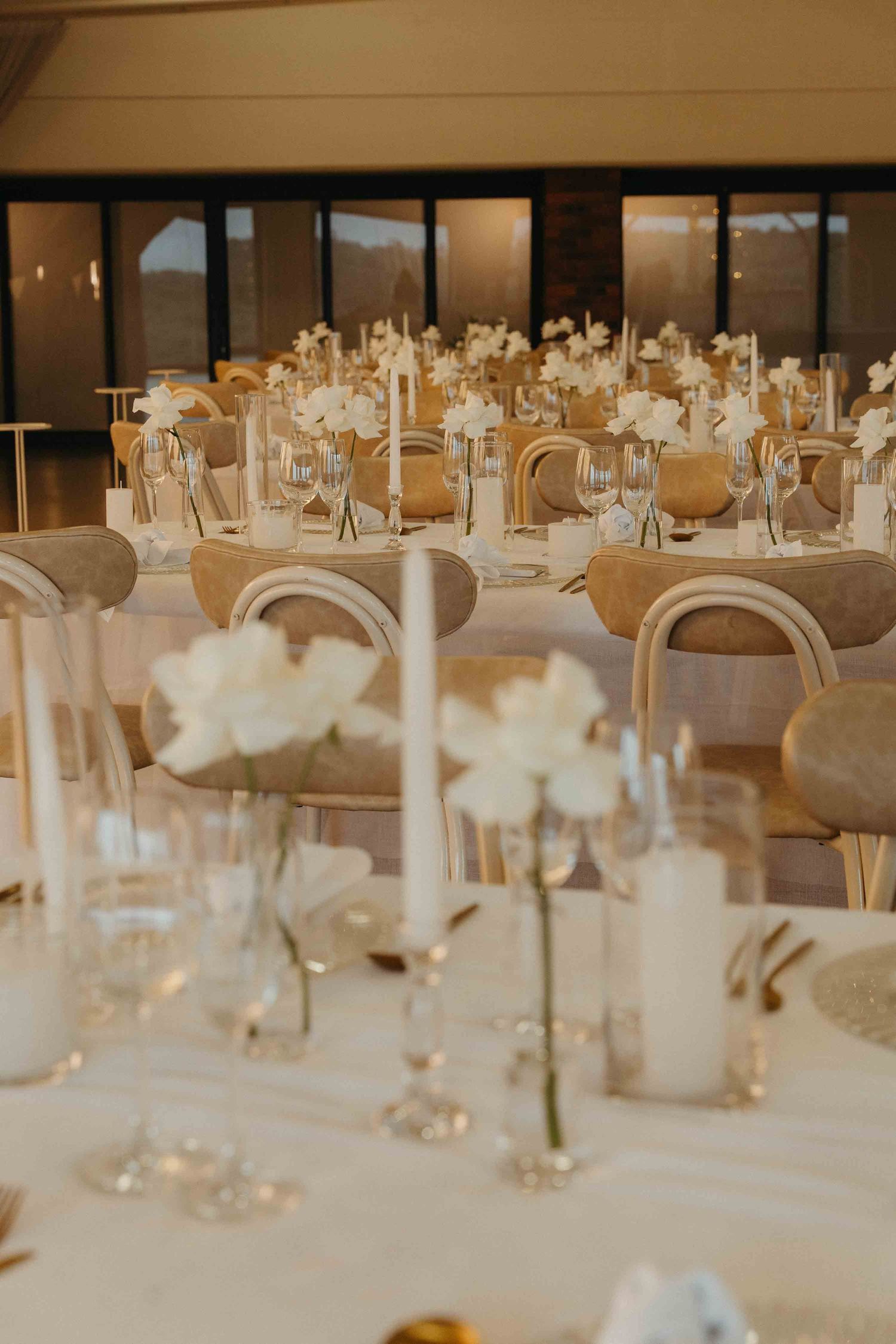 Elegant wedding reception tables with white floral centerpieces, tall candles, and neutral-toned chairs in a warmly lit venue.