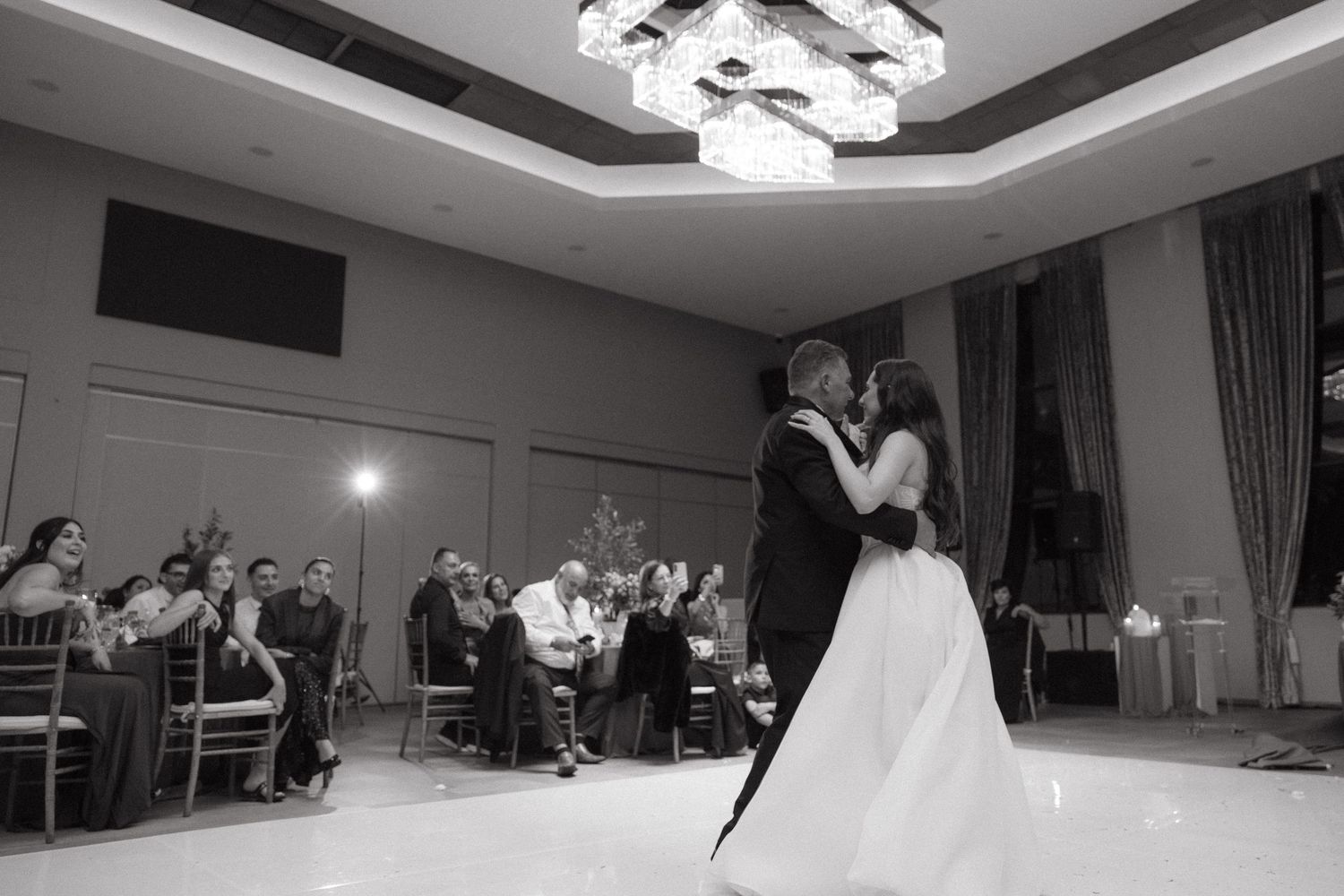 Black and white photo of bride and groom sharing first dance under chandelier with wedding guests seated in background.