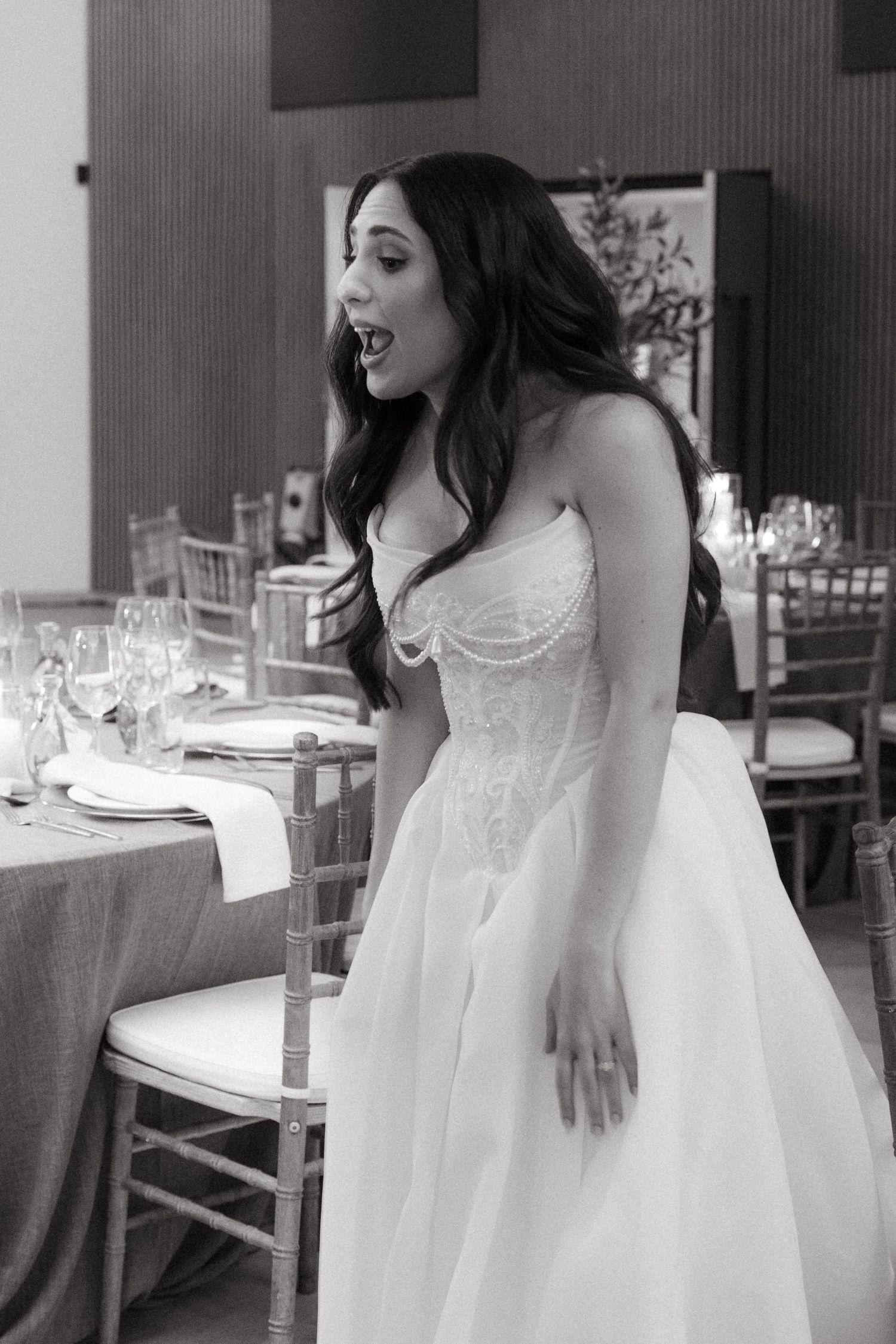 Bride in strapless lace ballgown reacts with surprise in elegant wedding reception venue, black and white photo.