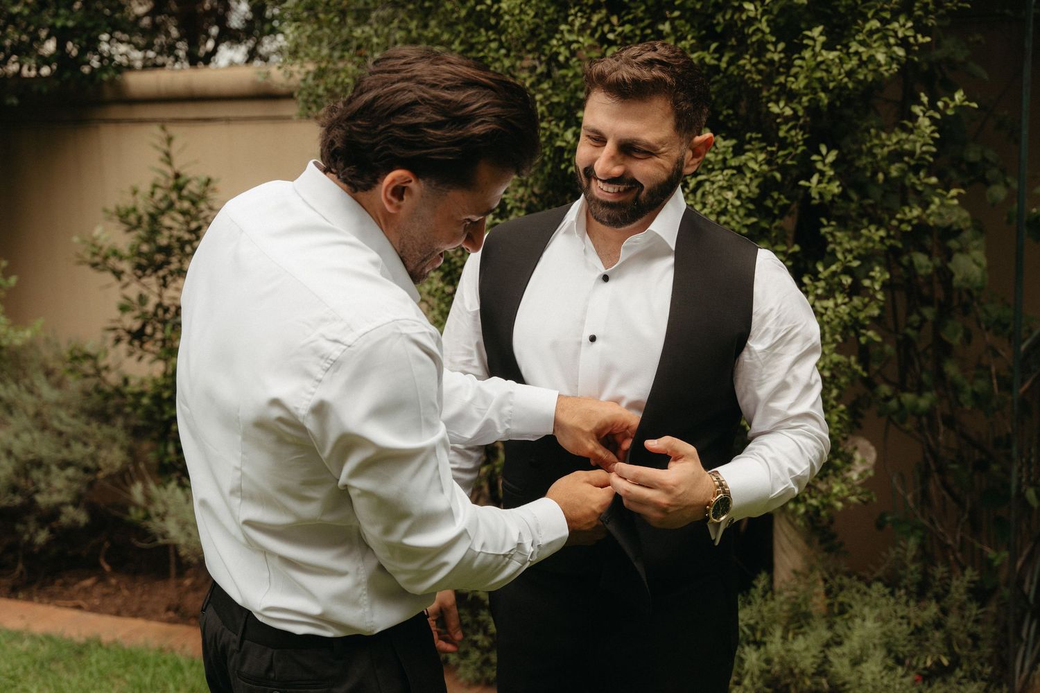 Two groomsmen in formal attire helping each other with cufflinks outdoors before a wedding ceremony.