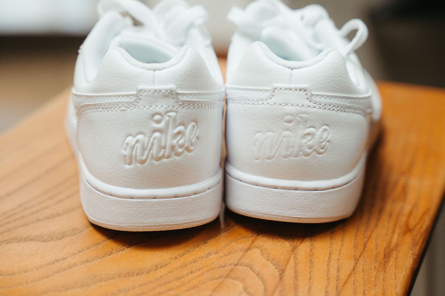 White Nike sneakers displayed heel-up on a wooden surface, showcasing embroidered Nike script logo.