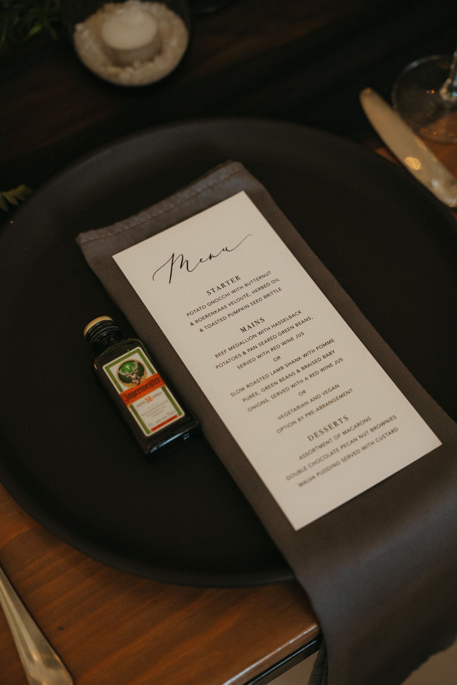 Elegant wedding menu card on black plate with dark napkin and mini Jagermeister bottle at formal dinner setting.