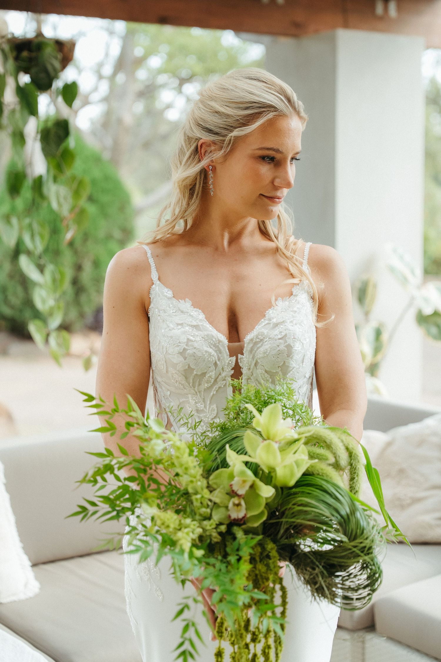 Blonde bride in lace wedding gown holding lush green tropical bouquet with orchids, standing in elegant outdoor venue.