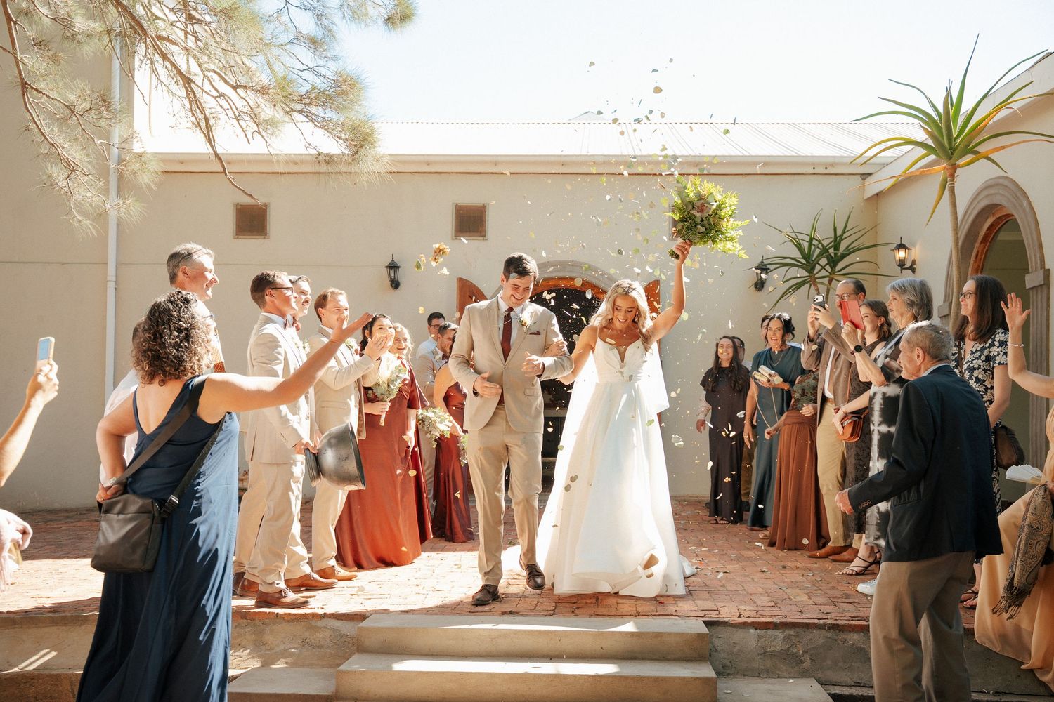 Bride and groom walk hand in hand with raised arms as joyful wedding guests cheer and celebrate outside a sunlit stucco venue.