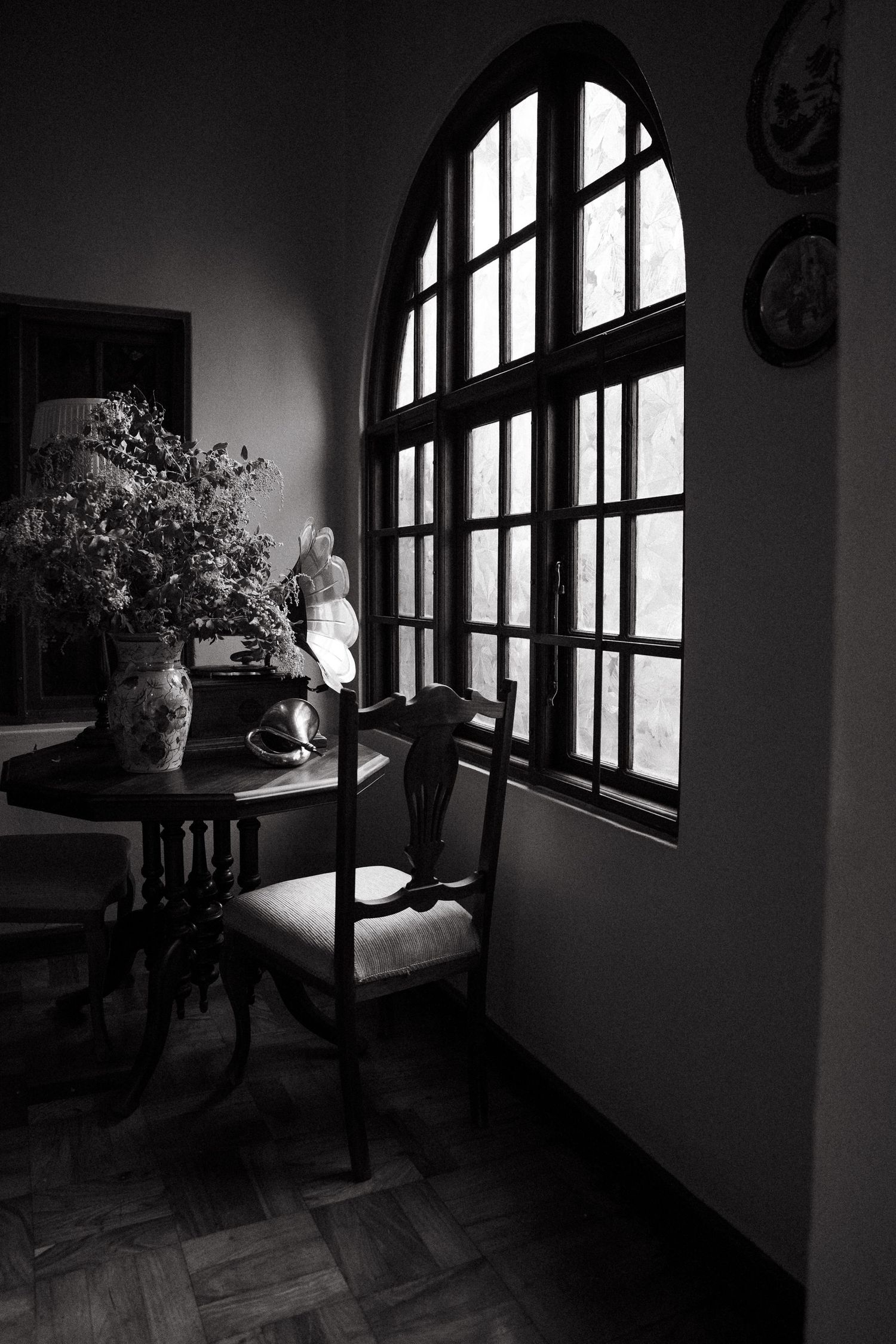 Black and white photo of elegant dining room with arched window, ornate wooden chair, round table with floral vase.