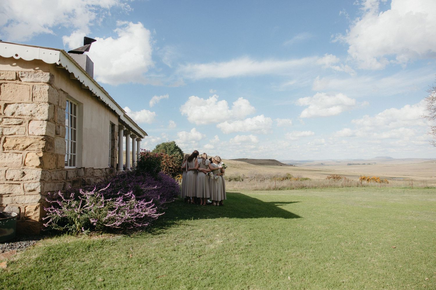 Bridesmaids in grey dresses stand beside a stone farmhouse under a blue sky with vast open grasslands in the background.