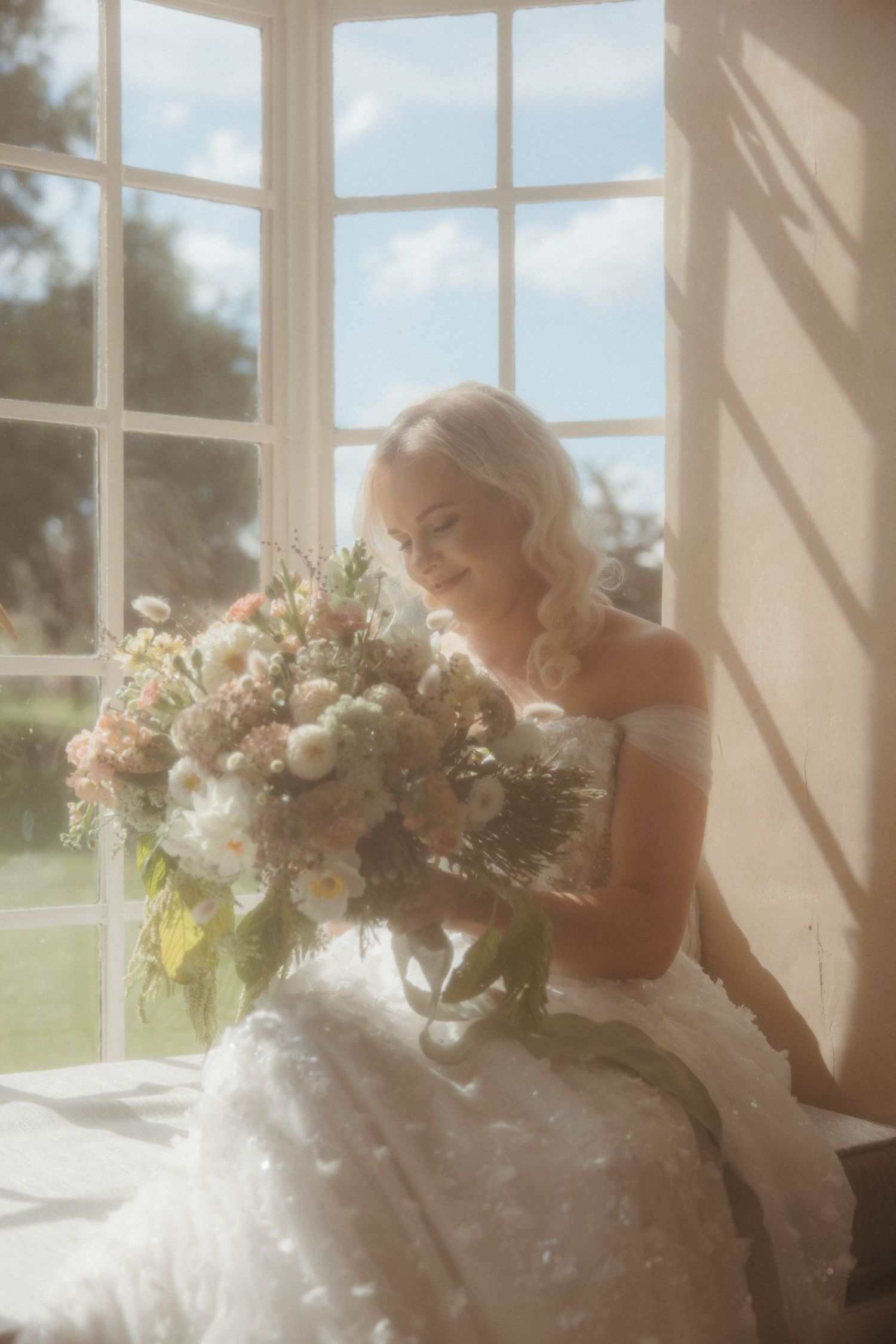 Bride in a white gown holds a lush bouquet by a sunlit window, smiling softly in a dreamy, ethereal setting.