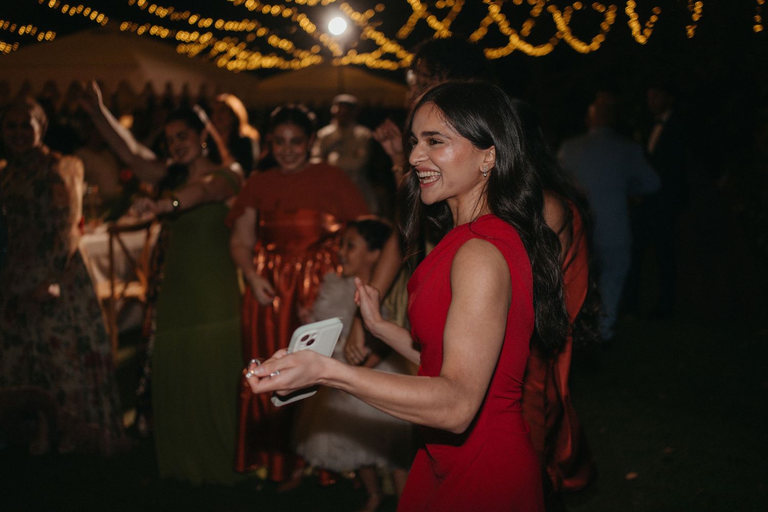 Woman in red dress smiling and clapping at an outdoor evening wedding reception with string lights.