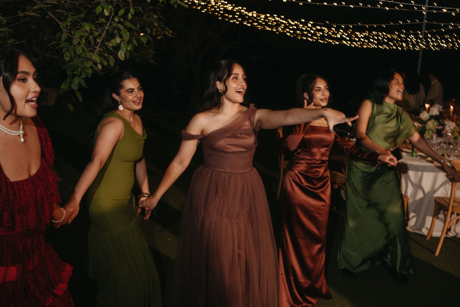 Five women in elegant gowns hold hands dancing outdoors at a wedding reception under string lights at night.