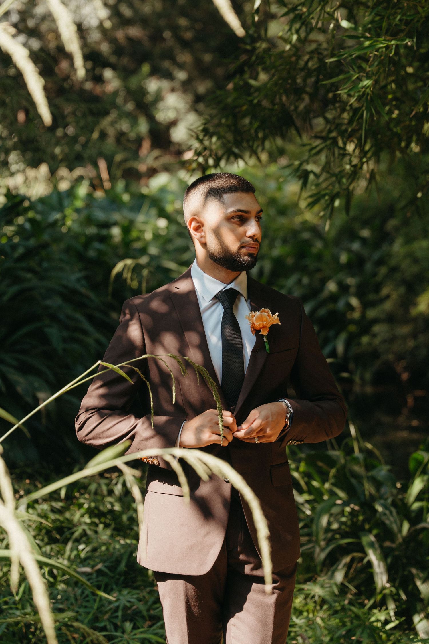 Groom in brown suit with orange boutonniere stands among lush green foliage in natural outdoor wedding portrait.